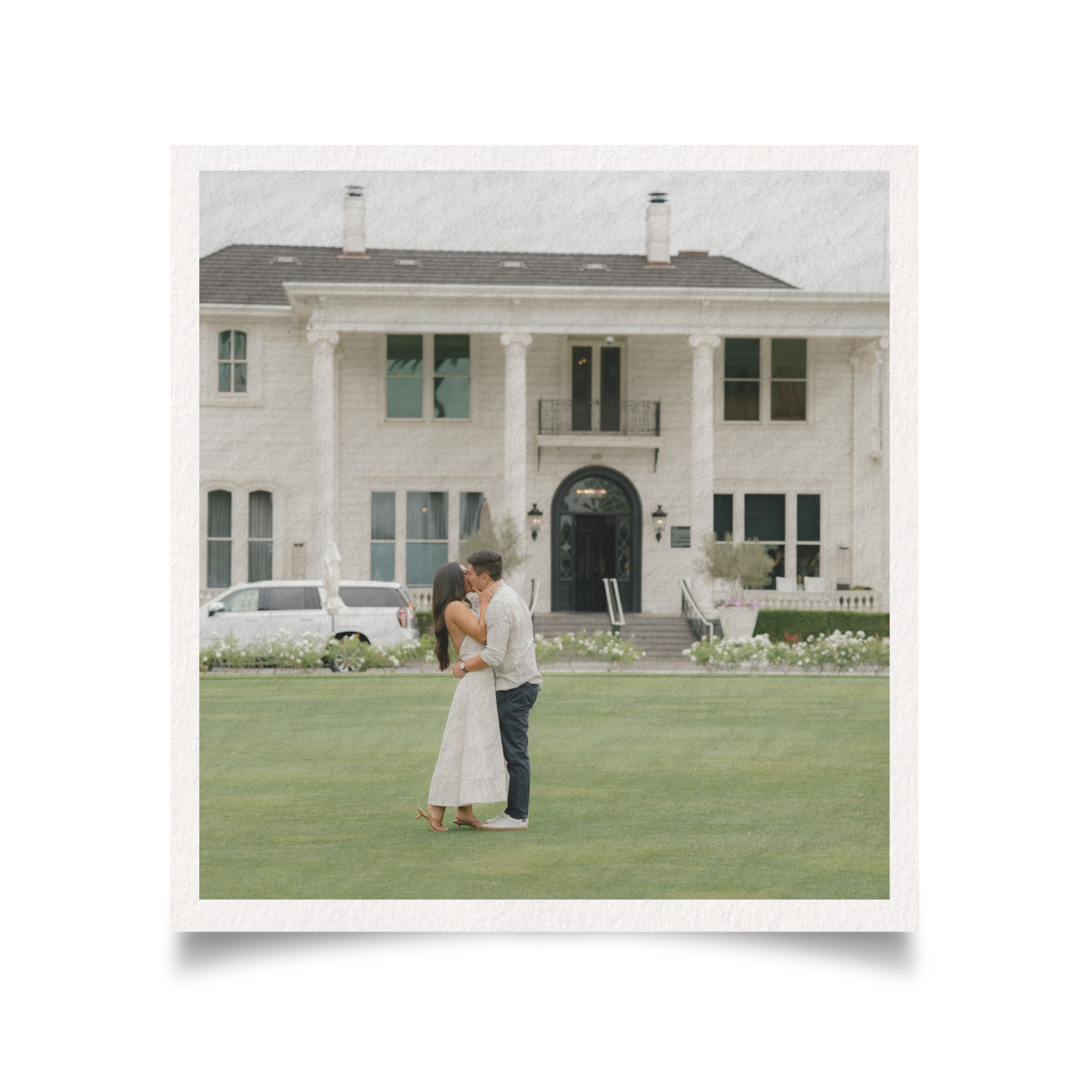 A couple sharing a kiss on a lawn in front of a large white house with columns and multiple windows.