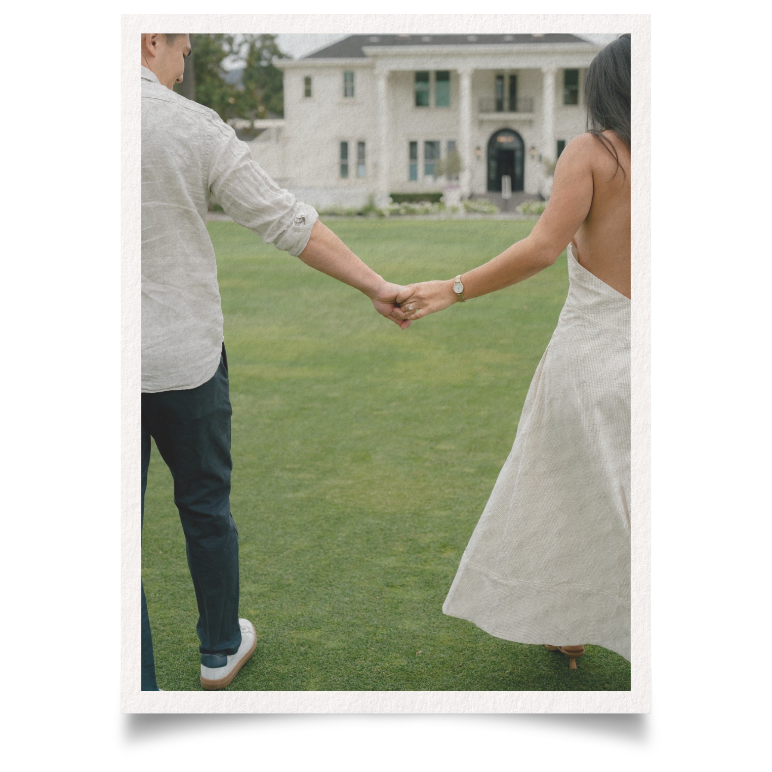 A couple holding hands and standing on a lawn in front of a large house, facing away from the camera.