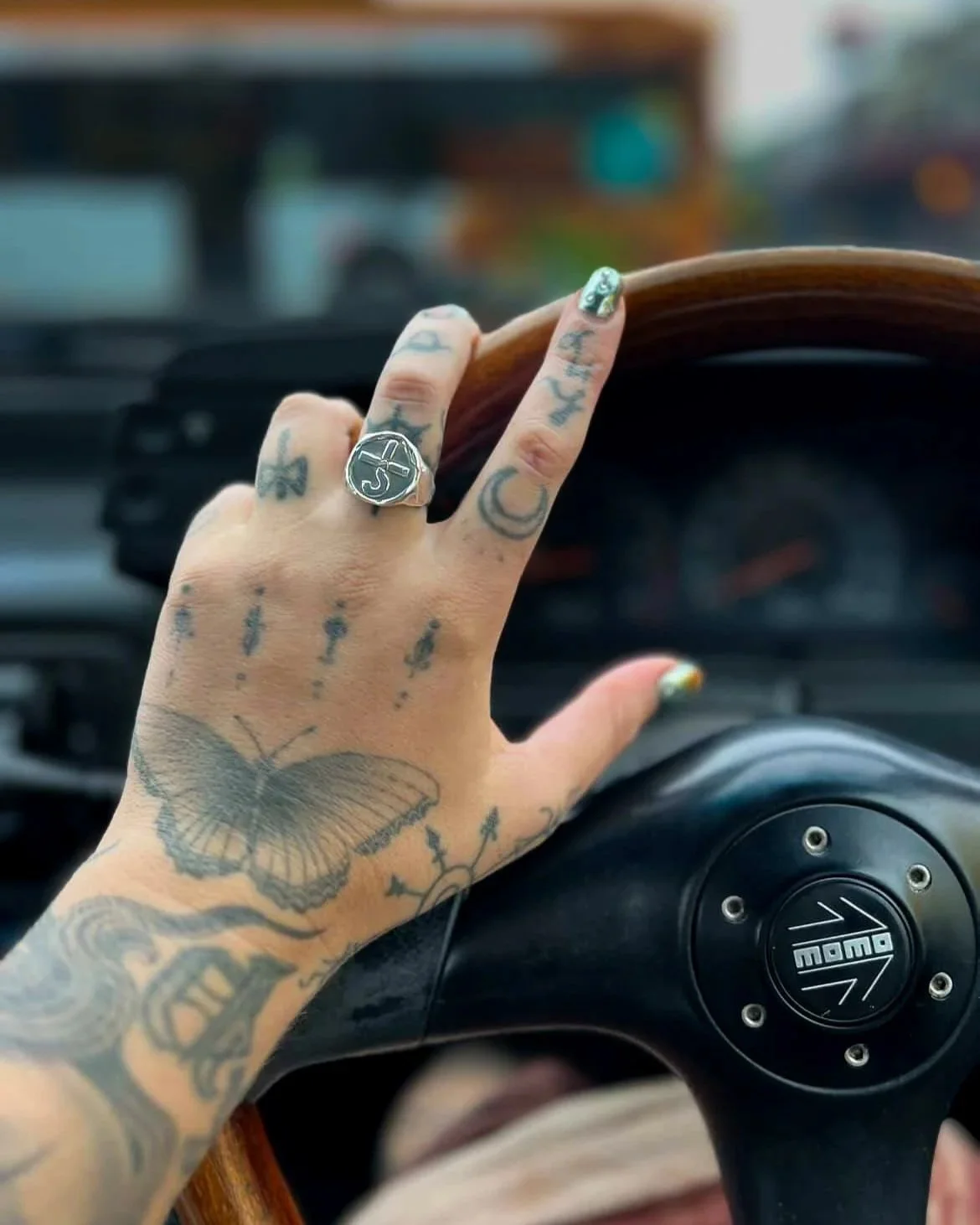 A person's hand with tattoos holding a wooden steering wheel inside a vehicle, wearing a signet ring with the blue oyster cult symbol on it.