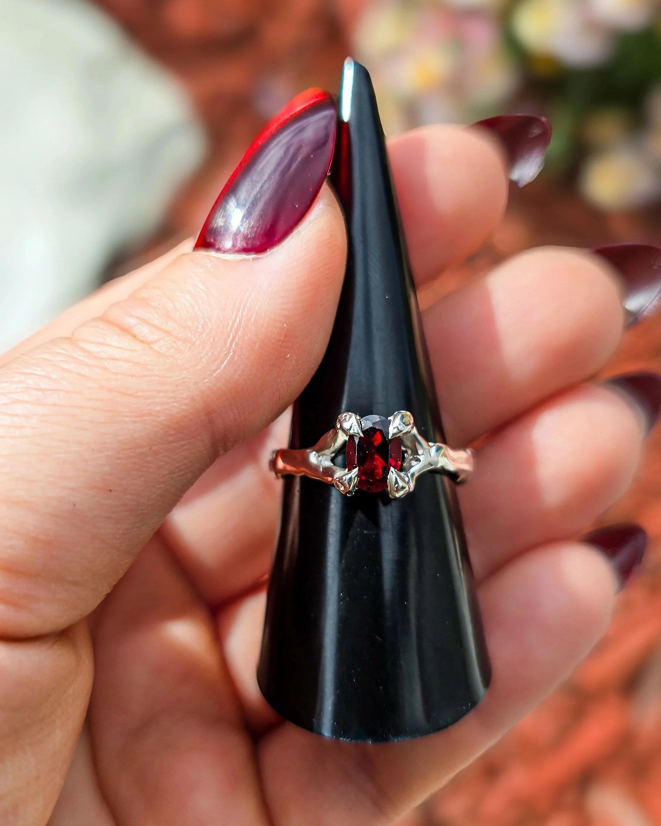 Close-up of a black ring holder with a silver ring that has a red garnet gemstone in a claw setting. Being held by a person's hand with matching red nail polish.