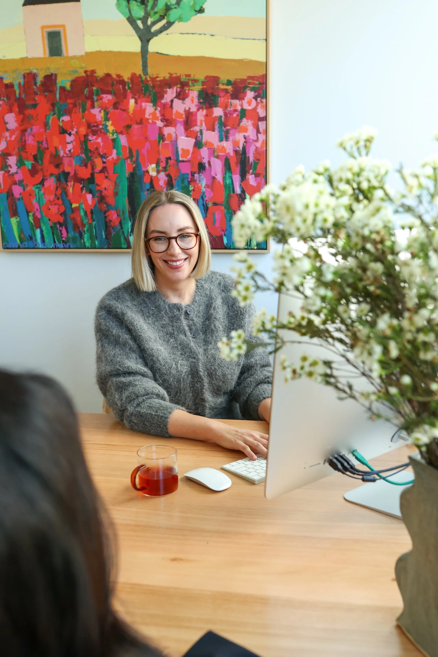 A woman with blonde hair and glasses smiling while sitting at a wooden desk with a large monitor, a white keyboard, a mouse, a glass mug with a dark drink, and a large bouquet of white flowers in the foreground. Behind her is a colorful painting of a landscape with a house, tree, and fields.