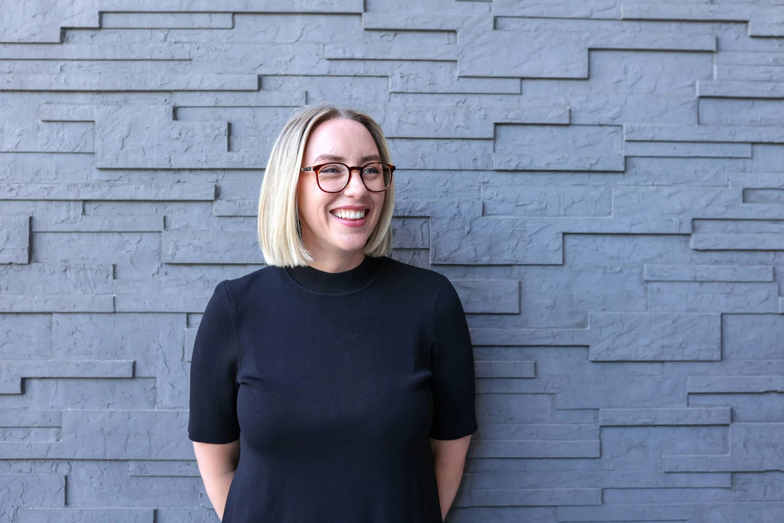 A woman with blonde hair, glasses, and a black top smiling and standing against a gray brick wall.