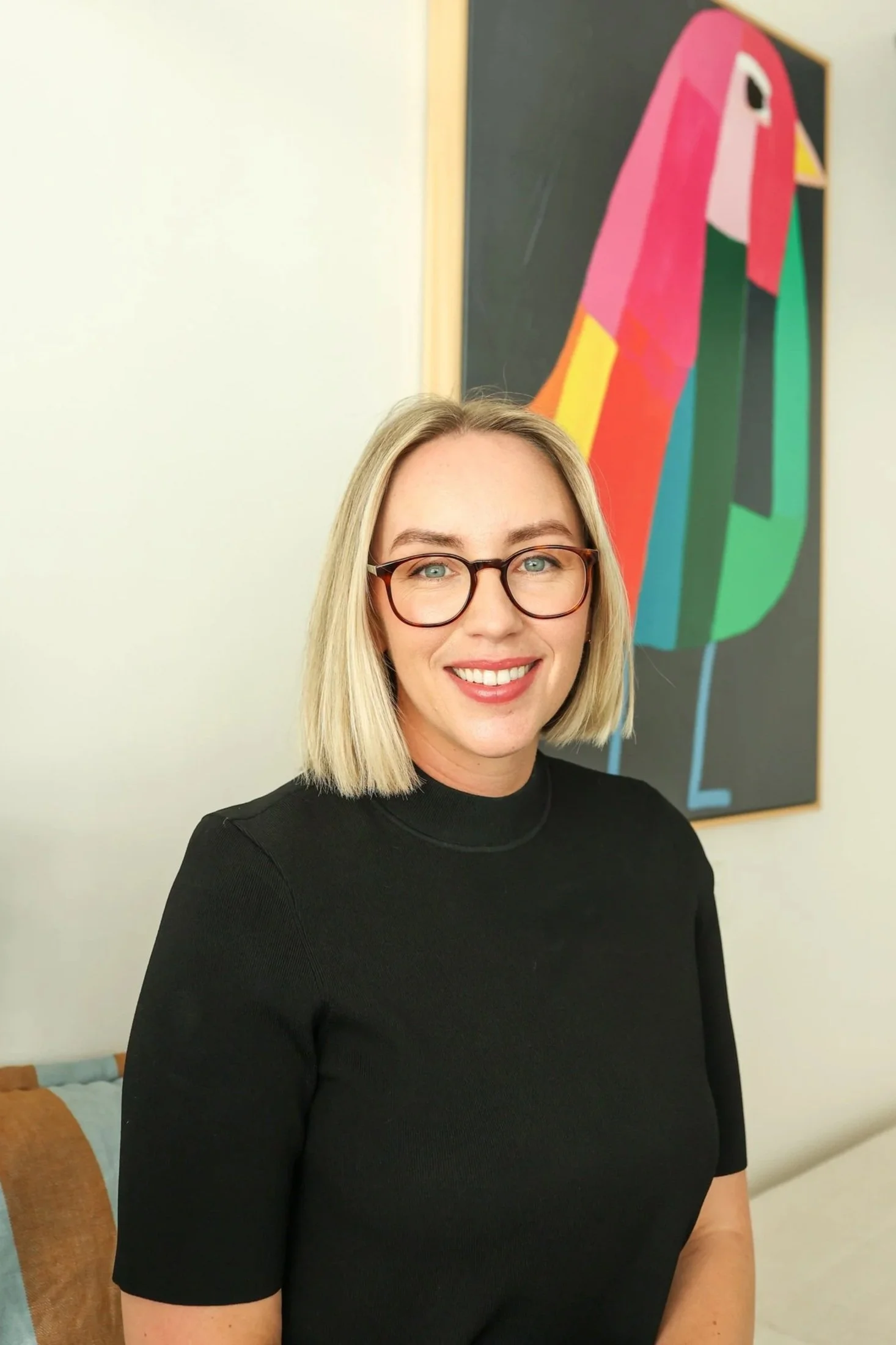 A woman with blonde hair, wearing glasses and a black shirt, smiling at the camera. Behind her is a colorful, geometric birds artwork on the wall.