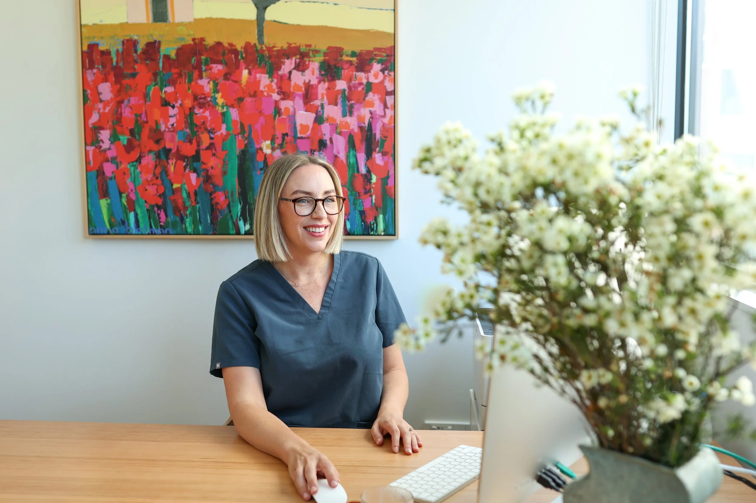A smiling woman wearing glasses and a navy medical uniform sitting at a desk with a computer mouse, keyboard, and a vase of white flowers, in a bright room with an abstract colorful painting on the wall.