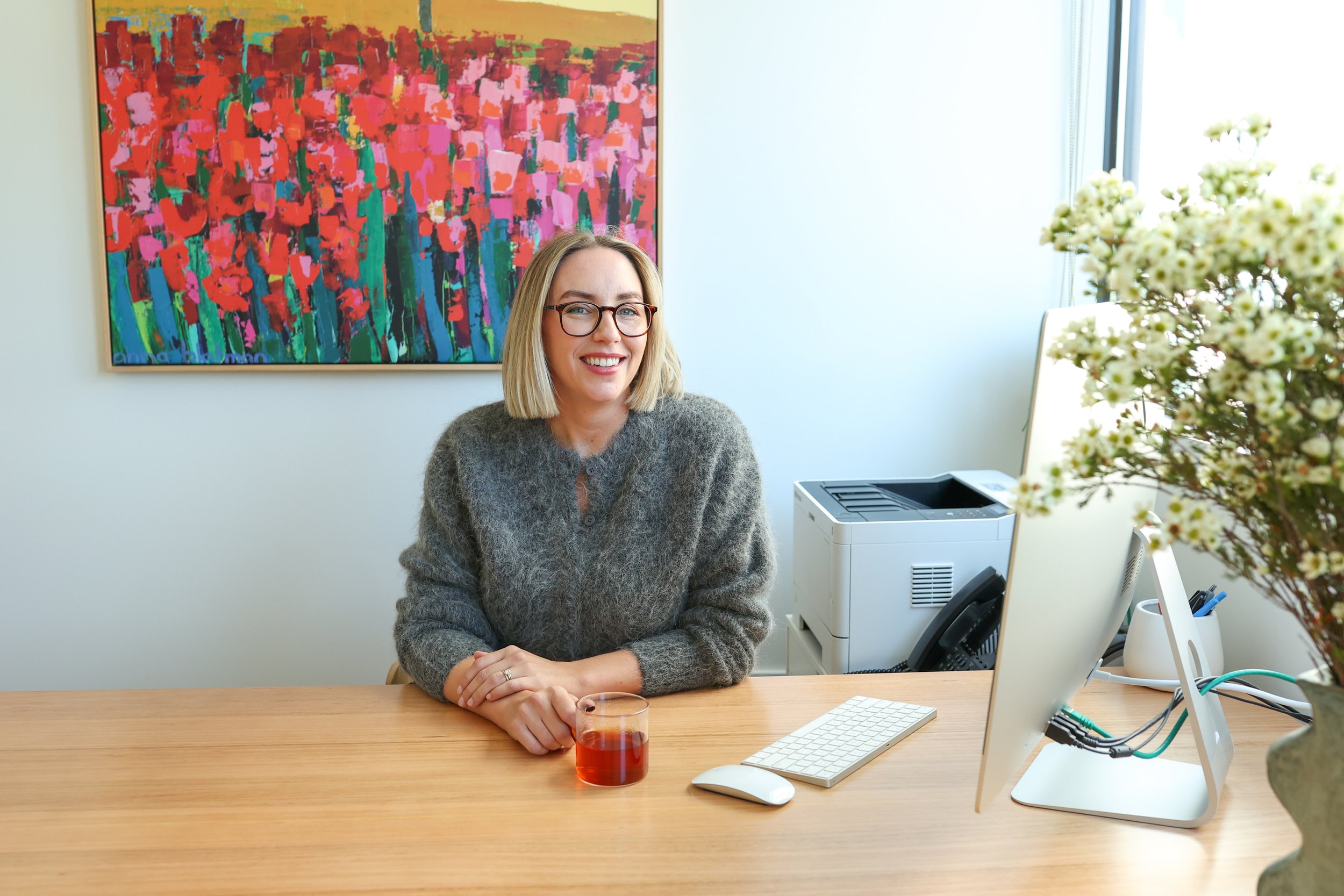 A woman with blonde hair, glasses, and a grey sweater sitting at a wooden desk in an office, smiling. There is a vase with white flowers, a computer monitor, keyboard, mouse, and a glass of red beverage on the desk. A colorful abstract painting hangs on the wall behind her, and a printer is on a stand beside her.