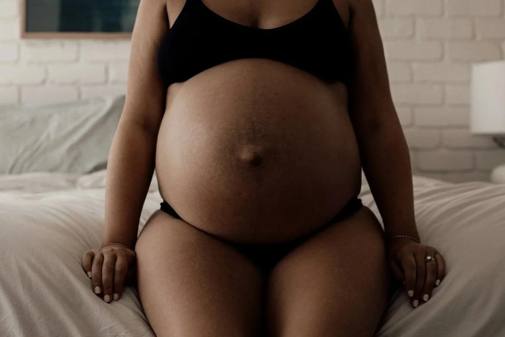 A pregnant woman wearing a black crop top and black underwear, sitting on a bed in a room with a white brick wall and a lamp.