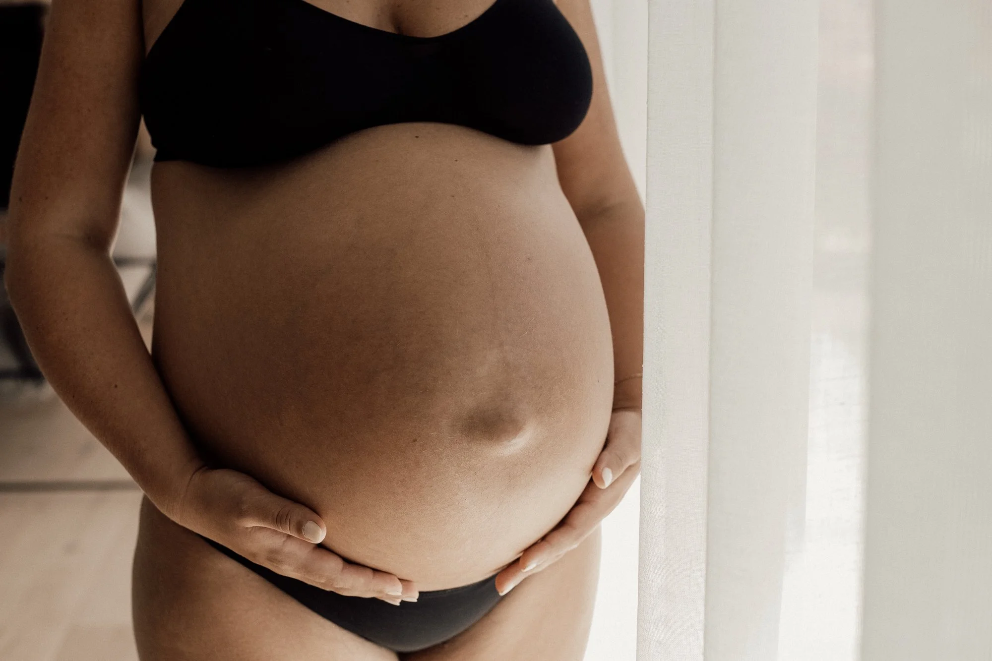 A pregnant woman stands near a window wearing a black sports bra and black underwear, with her hands resting on her belly.