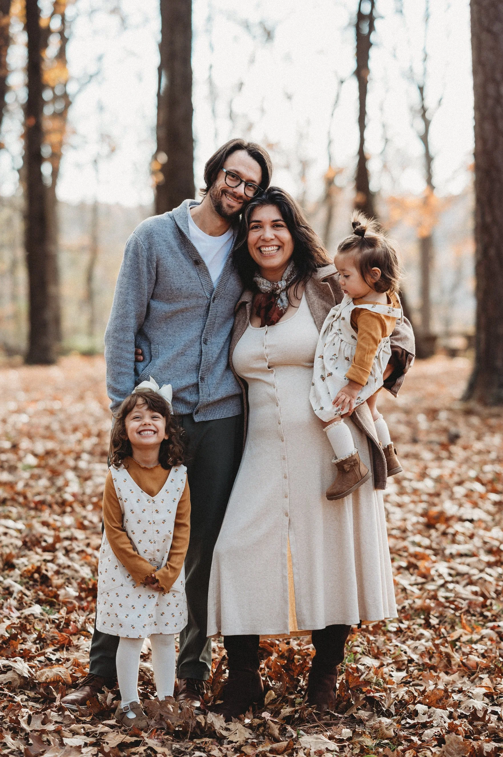 Family of four standing in a forest with autumn leaves, smiling and posing for the photo. The father has glasses and a gray sweater, the mother is wearing a long cream-colored dress, and the two young girls are dressed in fall clothing, one with a white dress and the other with a brown coat, all enjoying a fall day.