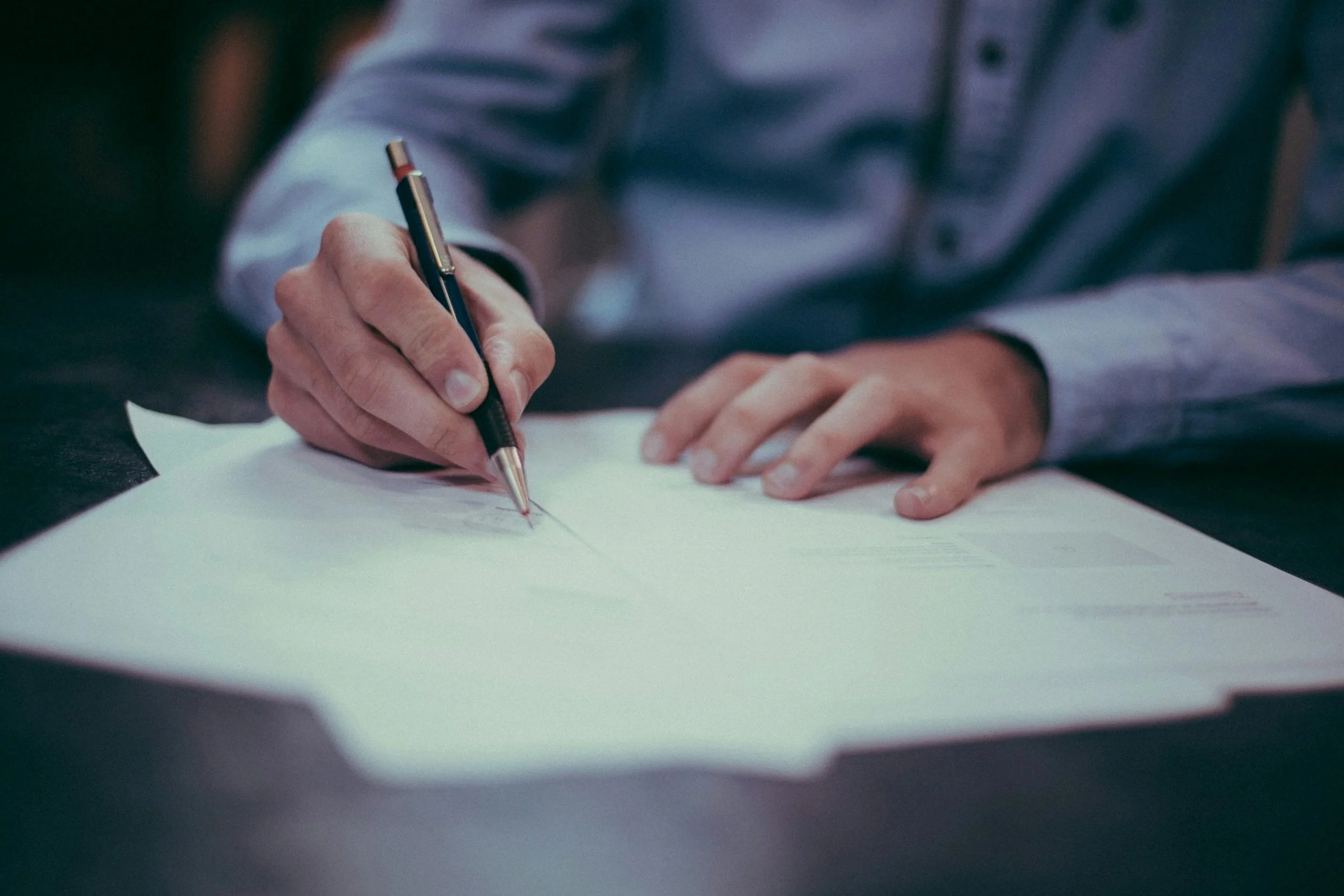 A person in a dress shirt is writing with a pen on a sheet of paper on a dark table.