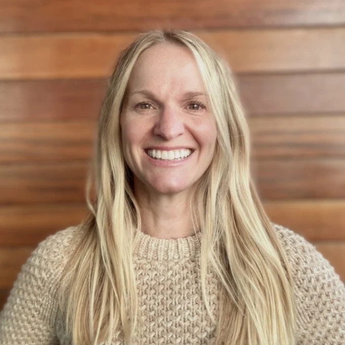 A smiling woman with long blonde hair wearing a beige knitted sweater against a wooden wall background.