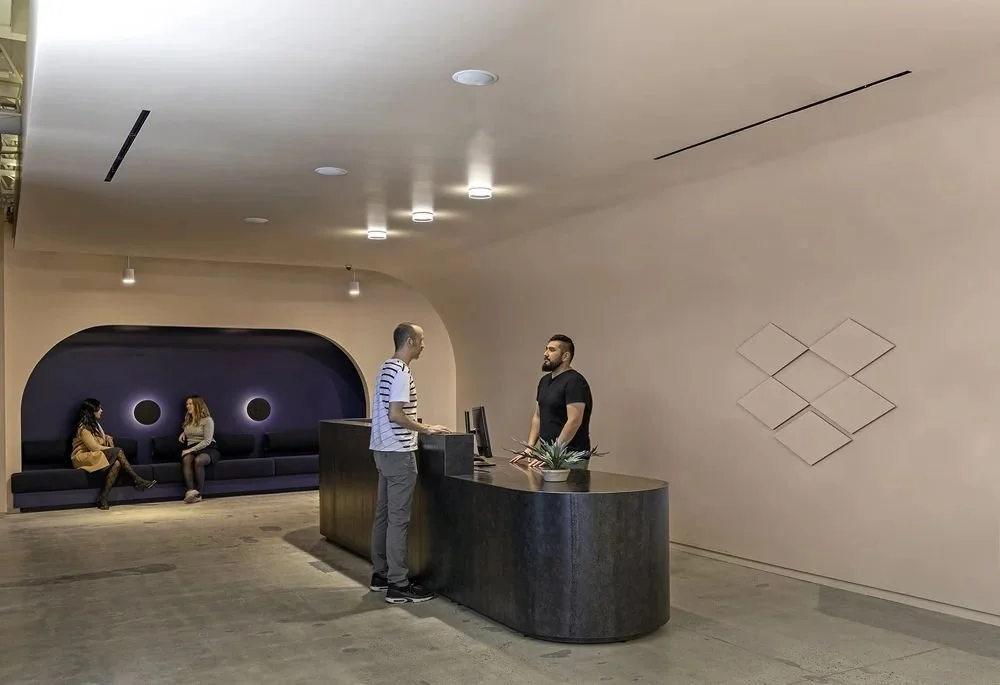 Modern hotel lobby reception area with a curved desk, two men checking in, and two women sitting on a black sofa in an arched alcove.