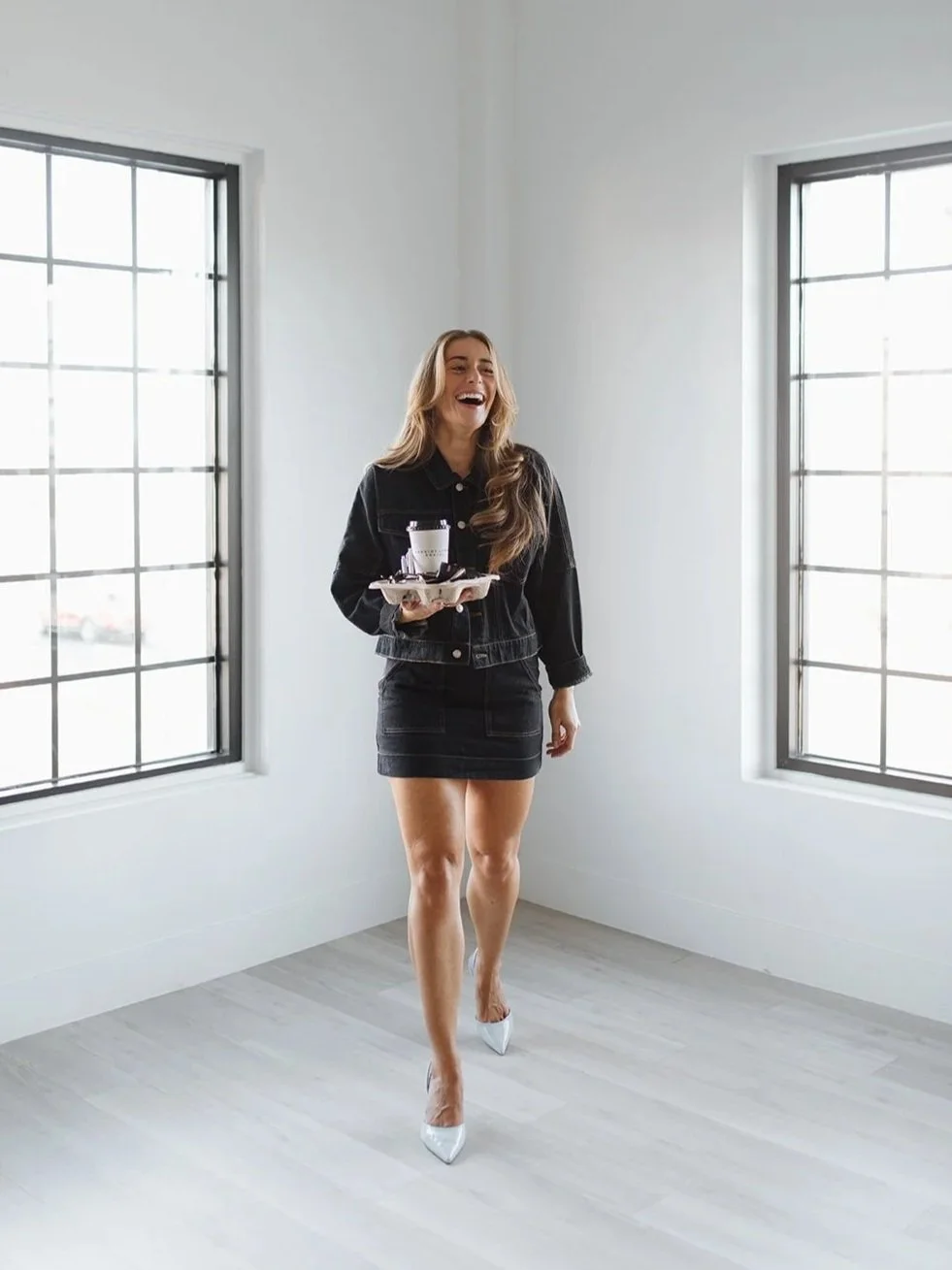 A woman in a black denim jacket and skirt walking in a bright room, laughing and holding a tray with a cup and a small plate.