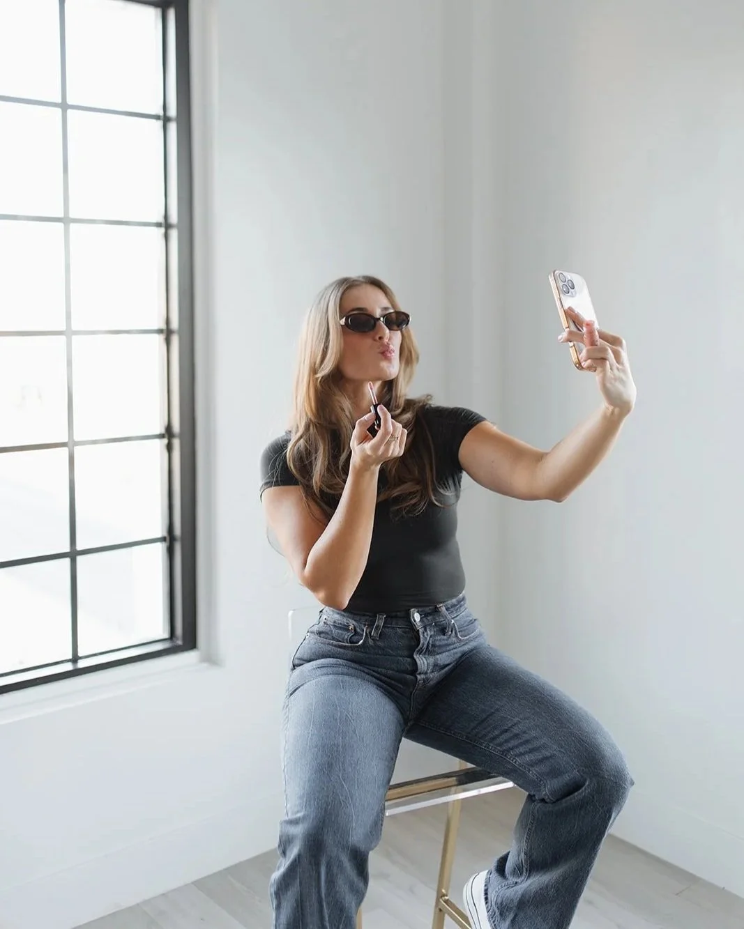 A woman in black shirt and jeans taking a selfie with her phone while applying lipstick near a window.
