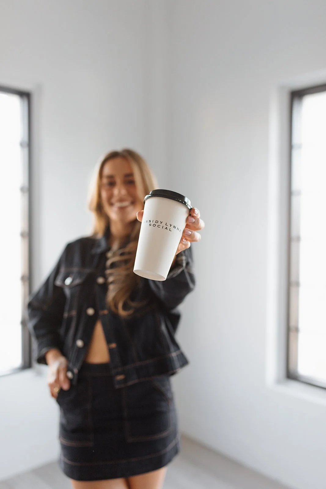 A woman with long hair smiling and holding a white disposable coffee cup towards the camera. She is wearing a black denim jacket and skirt, standing indoors with two windows in the background.