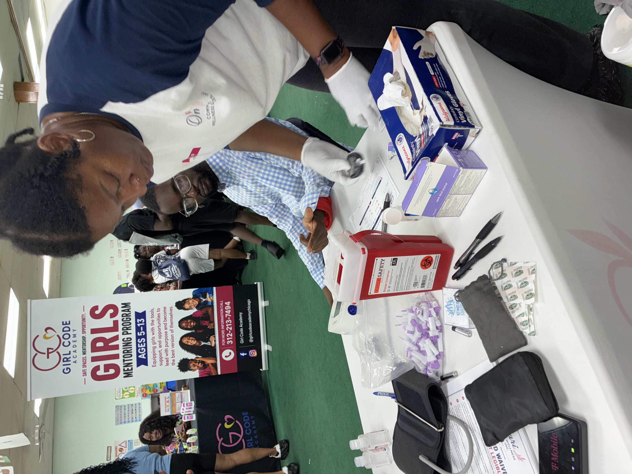 At a health awareness booth, two women are preparing to give a blood test with testing supplies on a table, including medical gloves, alcohol wipes, and a sharps disposal container.