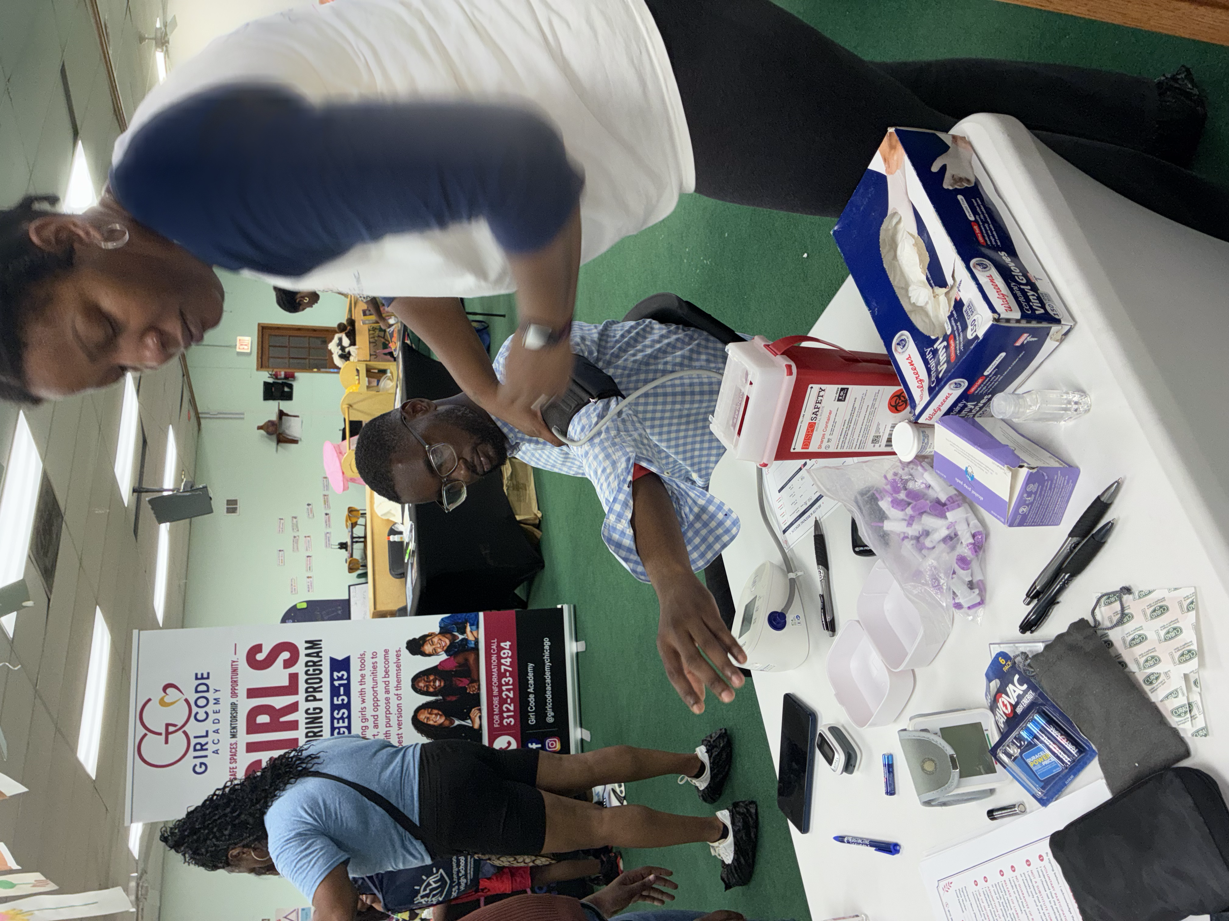 People gathered at a table with various health and safety items, including face masks, in a large room with banners and posters related to Girl Code Academy and girl empowerment.