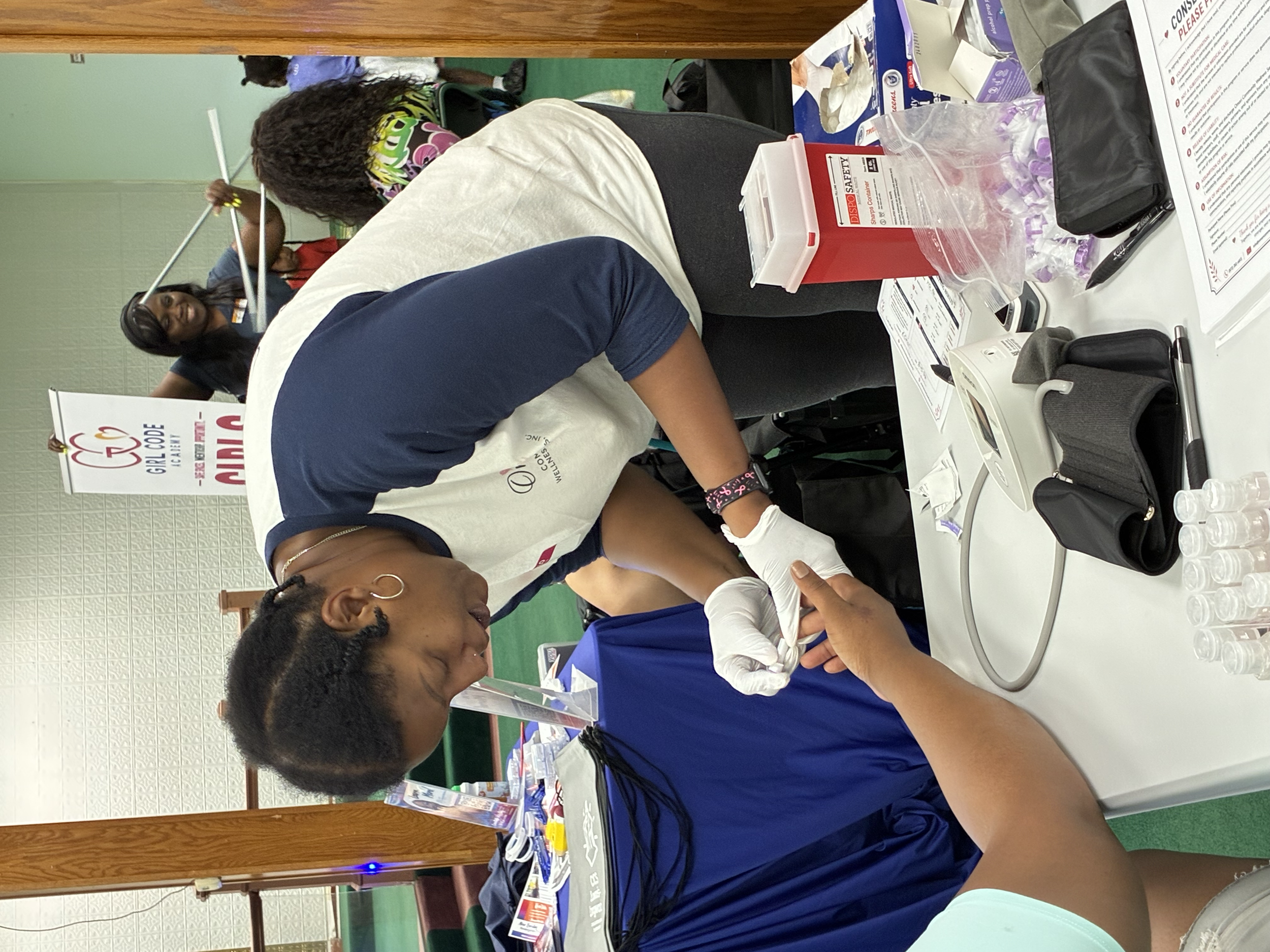 People participating in a blood donation event, with one woman donating blood at a table covered with medical supplies, and others seated nearby.
