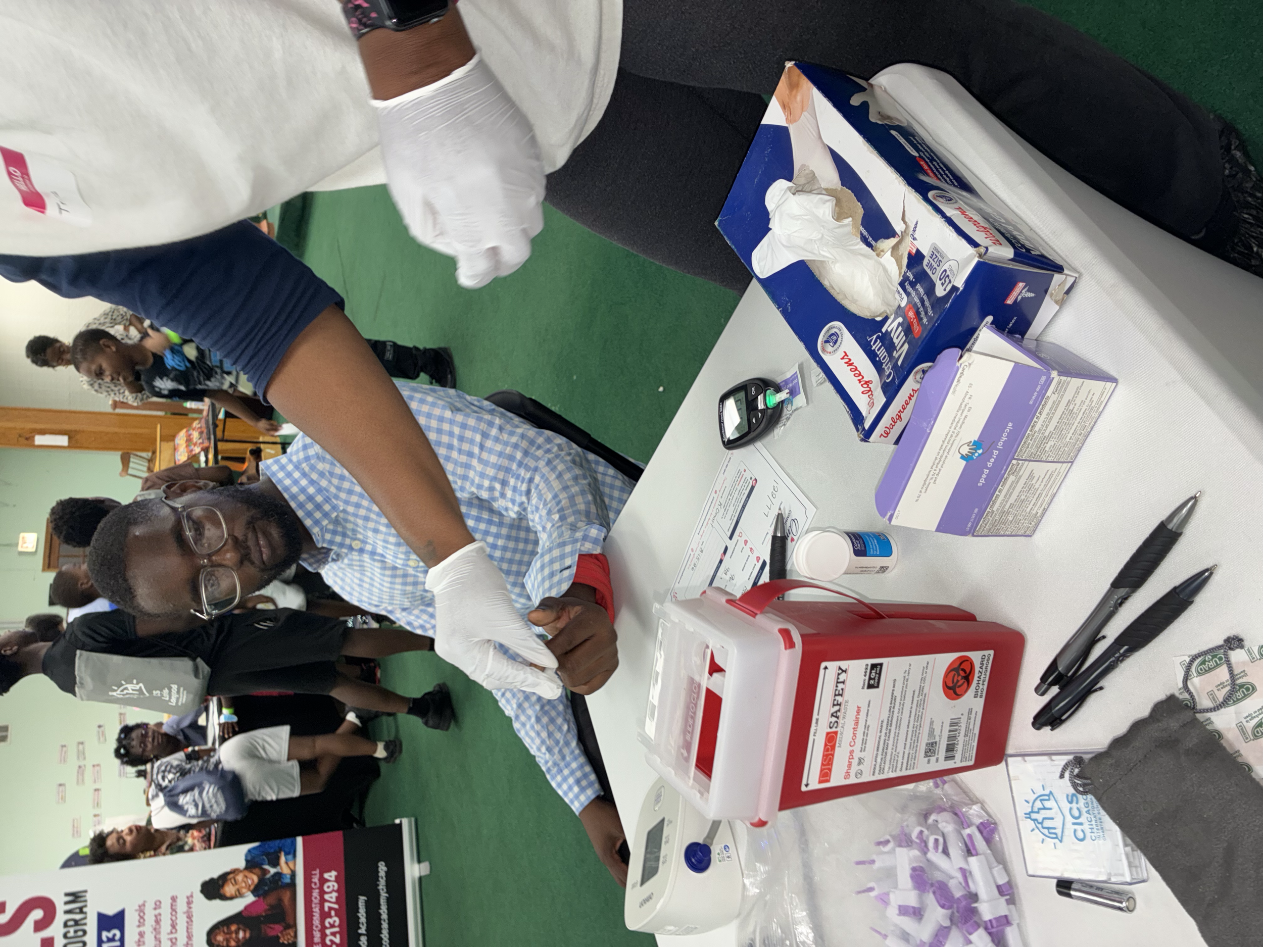 People gathered around a table at a health or safety event. The table has informational materials, including flyers, a blood pressure monitor, and health check supplies. In the background, there are more people and posters on the walls.