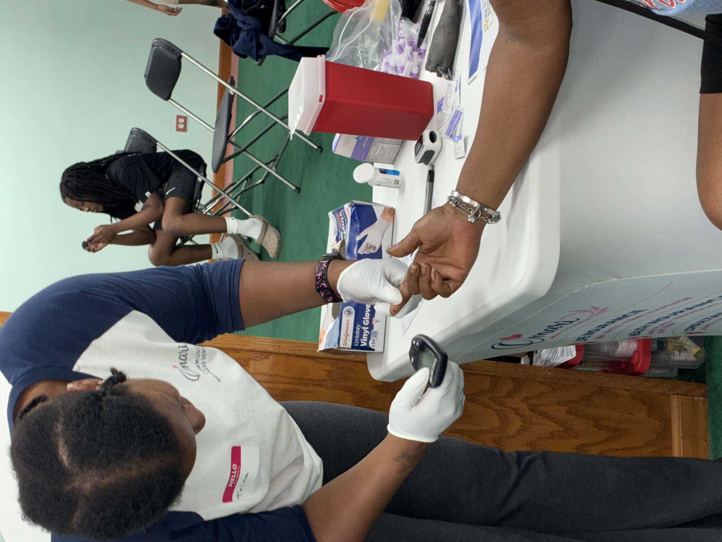 A person with dark curly hair getting a medical procedure on their arm, with another person holding their hand. The table has medical supplies including gloves, bandages, and a red container. There is another person sitting in the background.