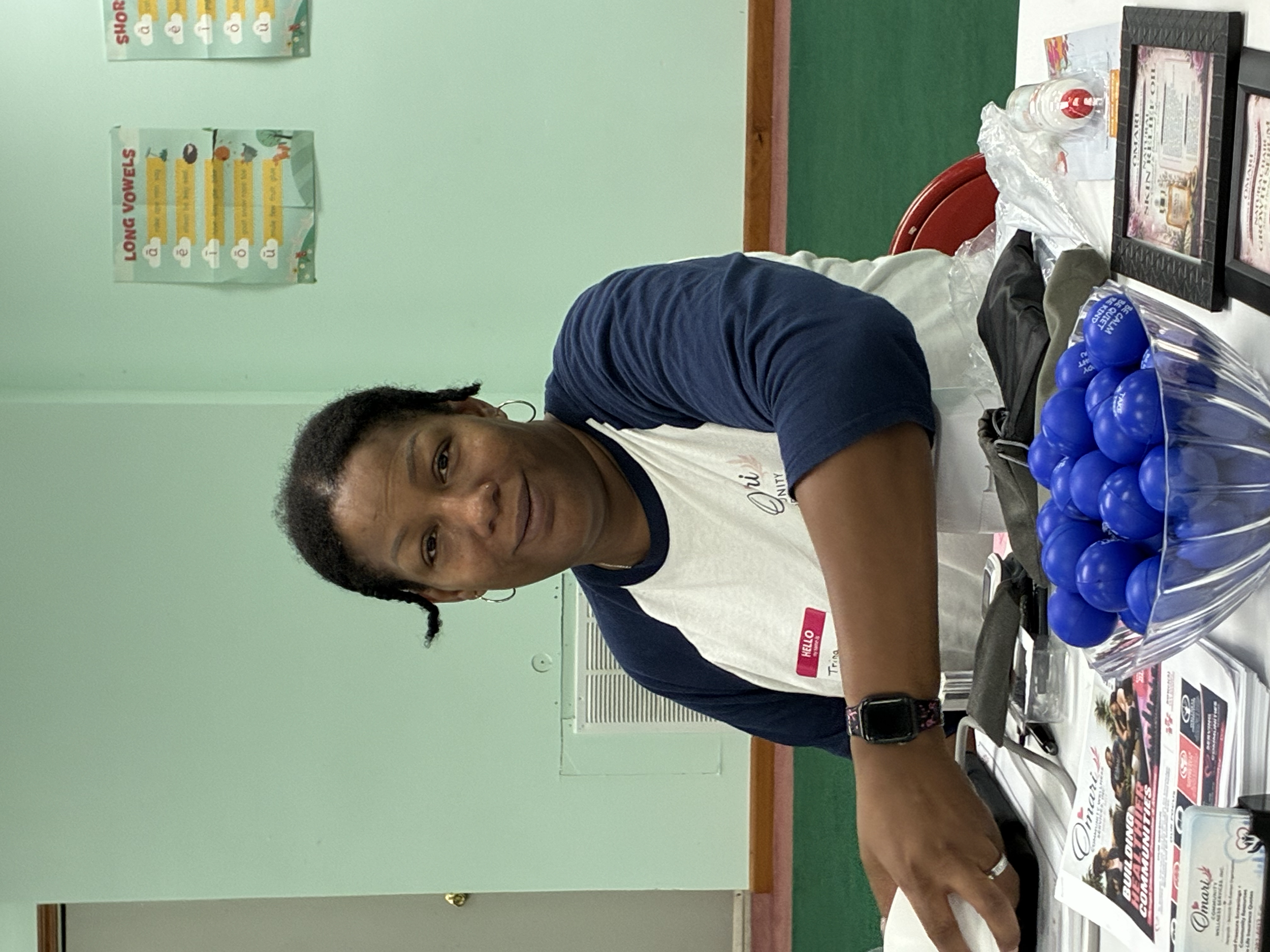 A woman with curly hair wearing a white and navy T-shirt stands beside a table with various magazines, flyers, and products, in a room with light green walls and a poster about long vowels on the wall.