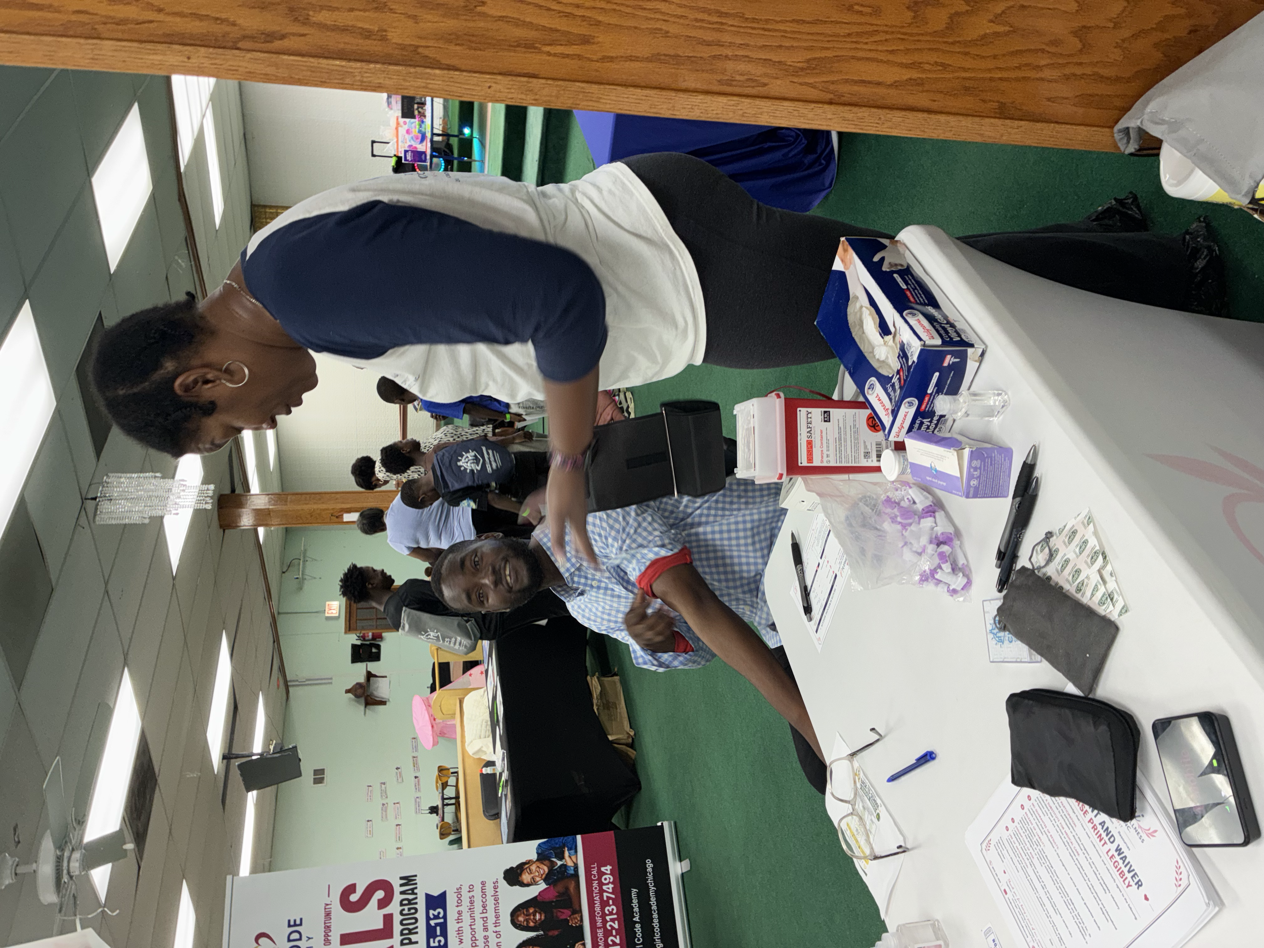 People standing and sitting at tables during an indoor event or conference, engaging in conversations and activities. Items on the table include papers, pens, and informational materials.
