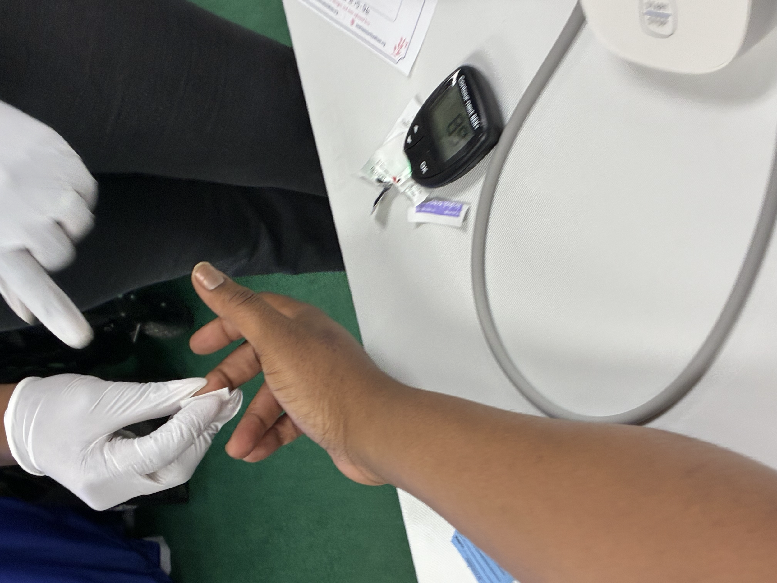 Two individuals, one in white gloves, are taking a blood sample from the arm of a person with medium skin tone at a medical setting. The scene occurs on a white table with medical supplies visible, including a blood glucose meter and paperwork.