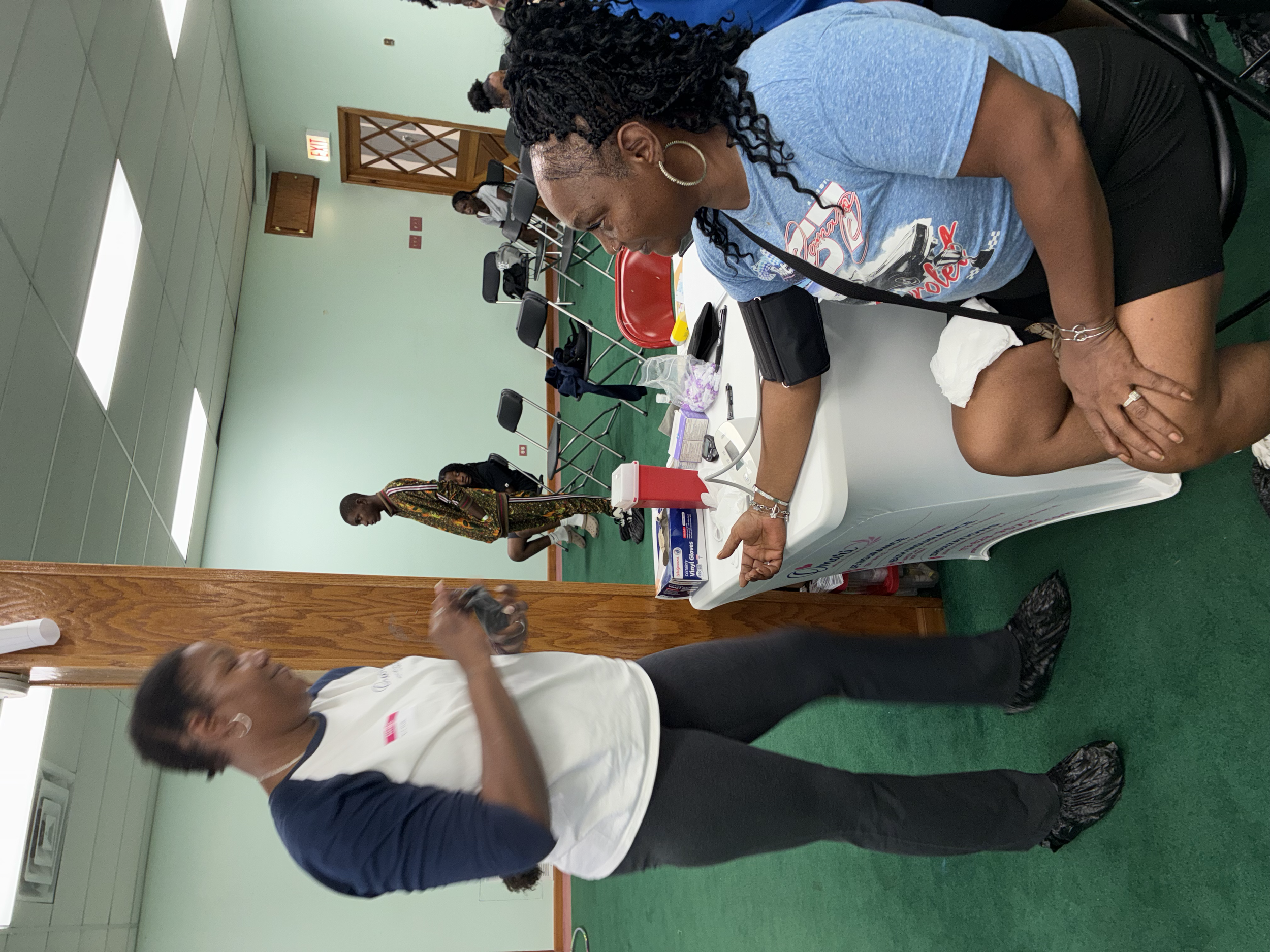 A woman with curly black hair, wearing a blue T-shirt, gold hoop earrings, and a silver bracelet, is sitting at a table in a room with mint green walls and a ceiling with fluorescent lights. She is looking down at her arm, possibly receiving an injec
