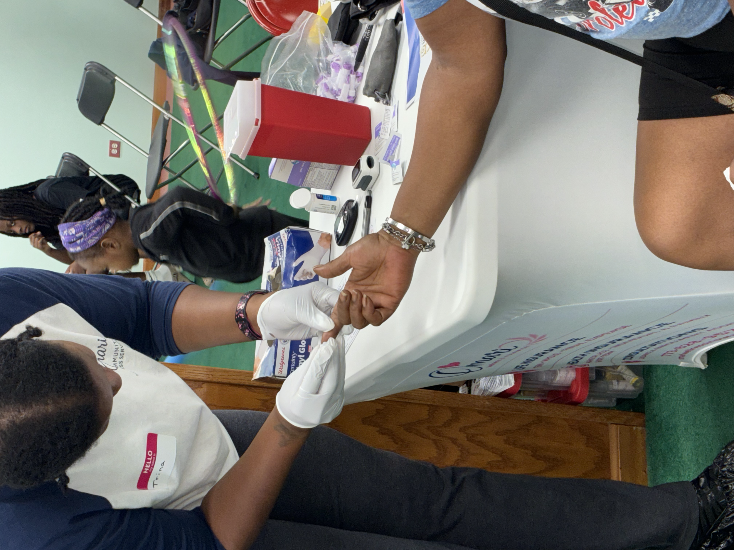 Medical setting with a patient seated at a table, receiving an injection or blood draw from a healthcare professional. The professional is wearing latex gloves and the patient has a band around their arm, possibly for a blood test. Medical supplies, 