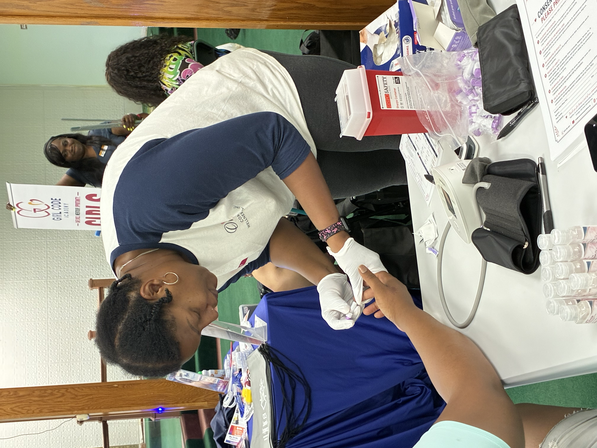 A group of people at a health or community event. Four women are visible, two seated and two standing. The seated women are receiving injections or vaccinations, handled by healthcare workers. The scene includes medical supplies on the table, promoti