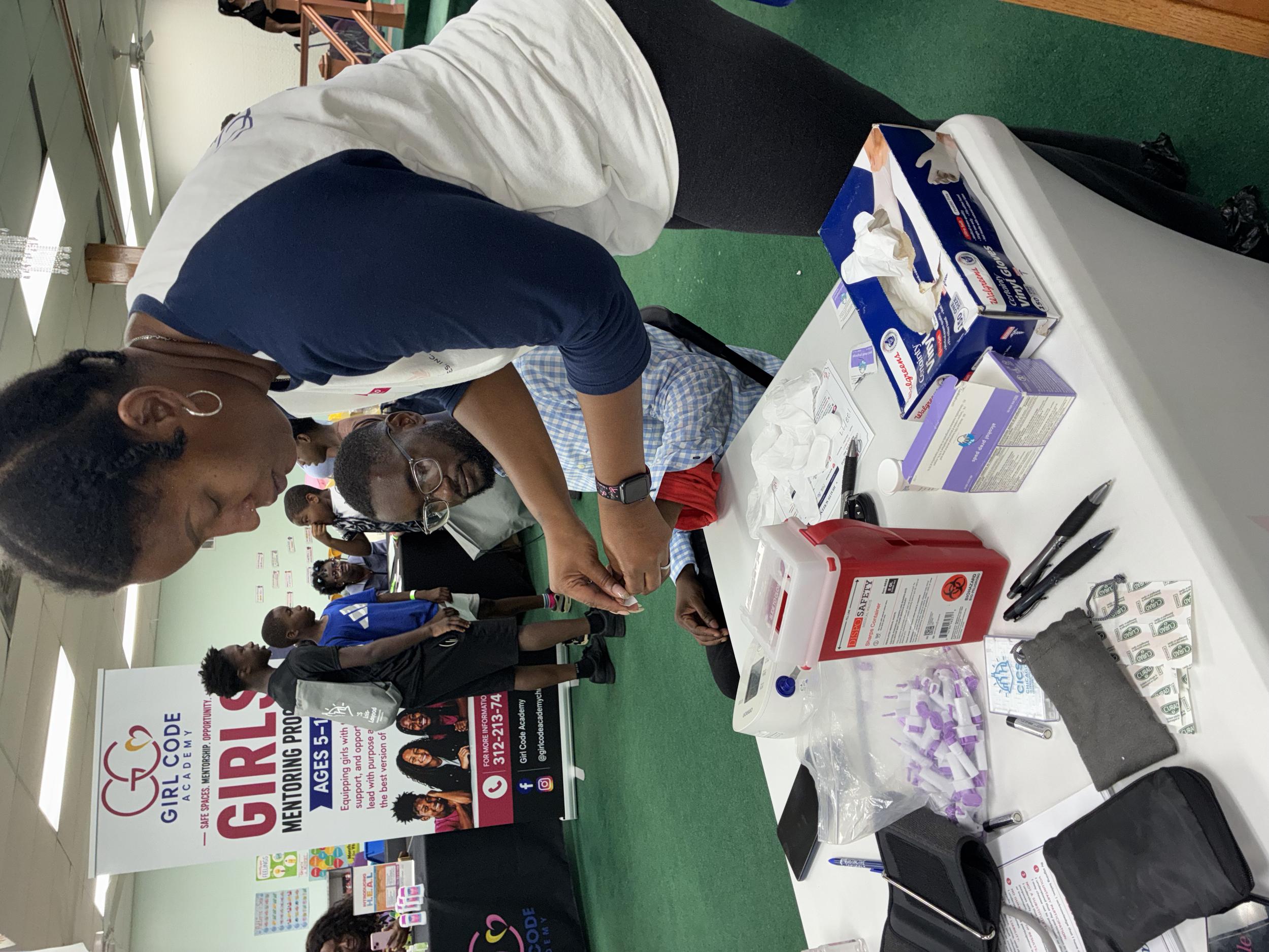 A table at a Girl Code Academy event with informational and promotional materials, including a box of tissues, pens, and pamphlets. People are gathered around the table, engaging and talking. A banner in the background features the Girl Code Academy 