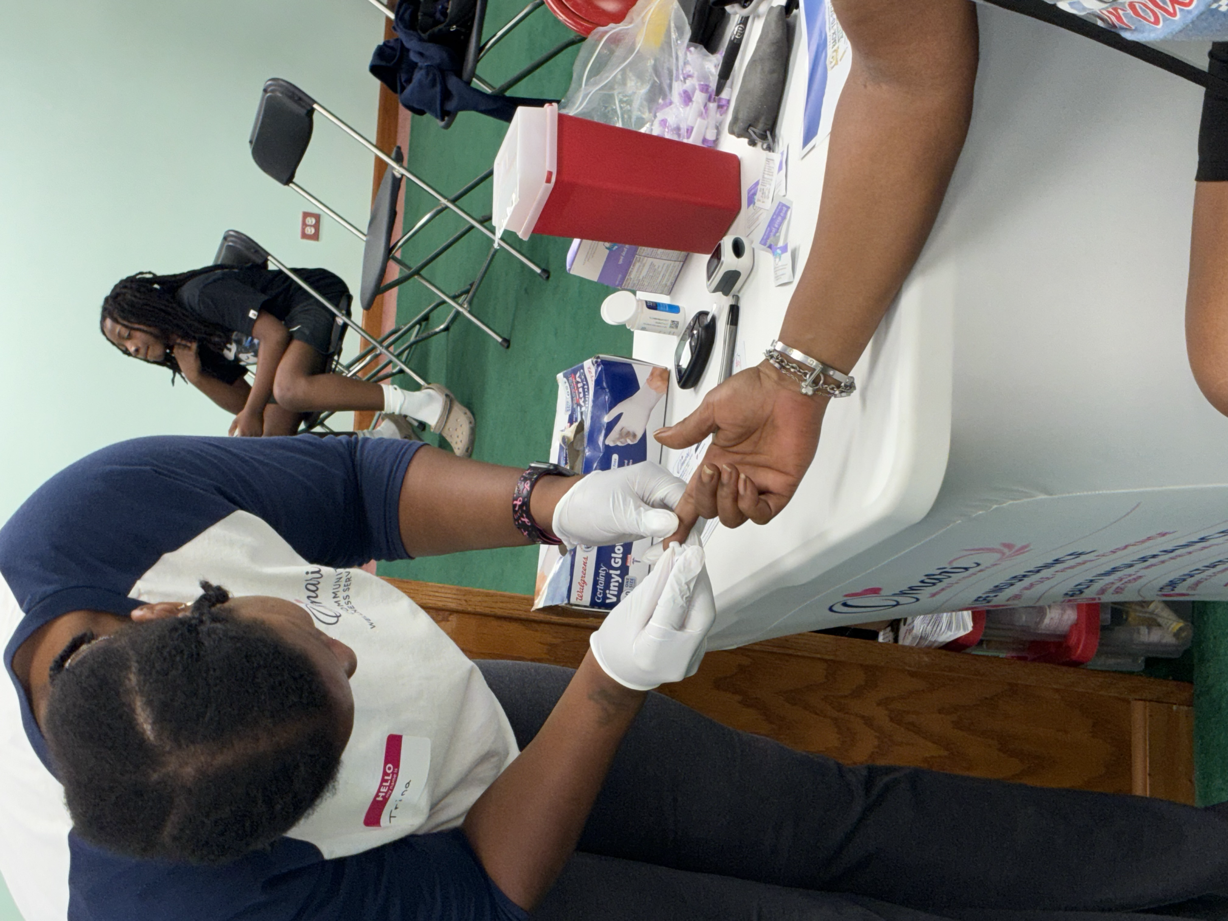 A person wearing a white shirt and white gloves is drawing blood from a person with dark skin and braided hair. The person donating blood is sitting at a table with various medical supplies and informational pamphlets on it. A young woman in a black 