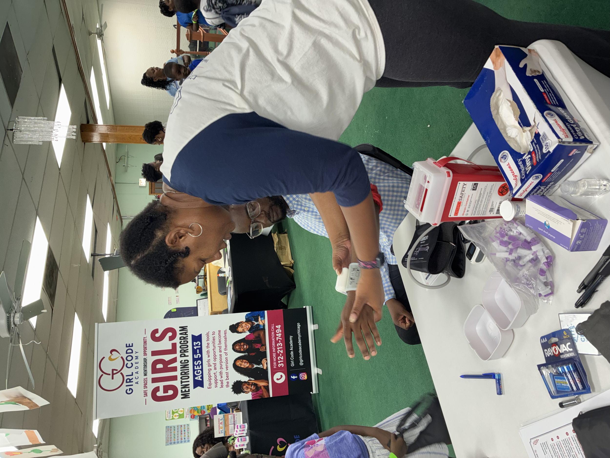 People at an event booth with a sign promoting a girls' mentorship program, visible health and safety items on the table, and several attendees engaging with the display.