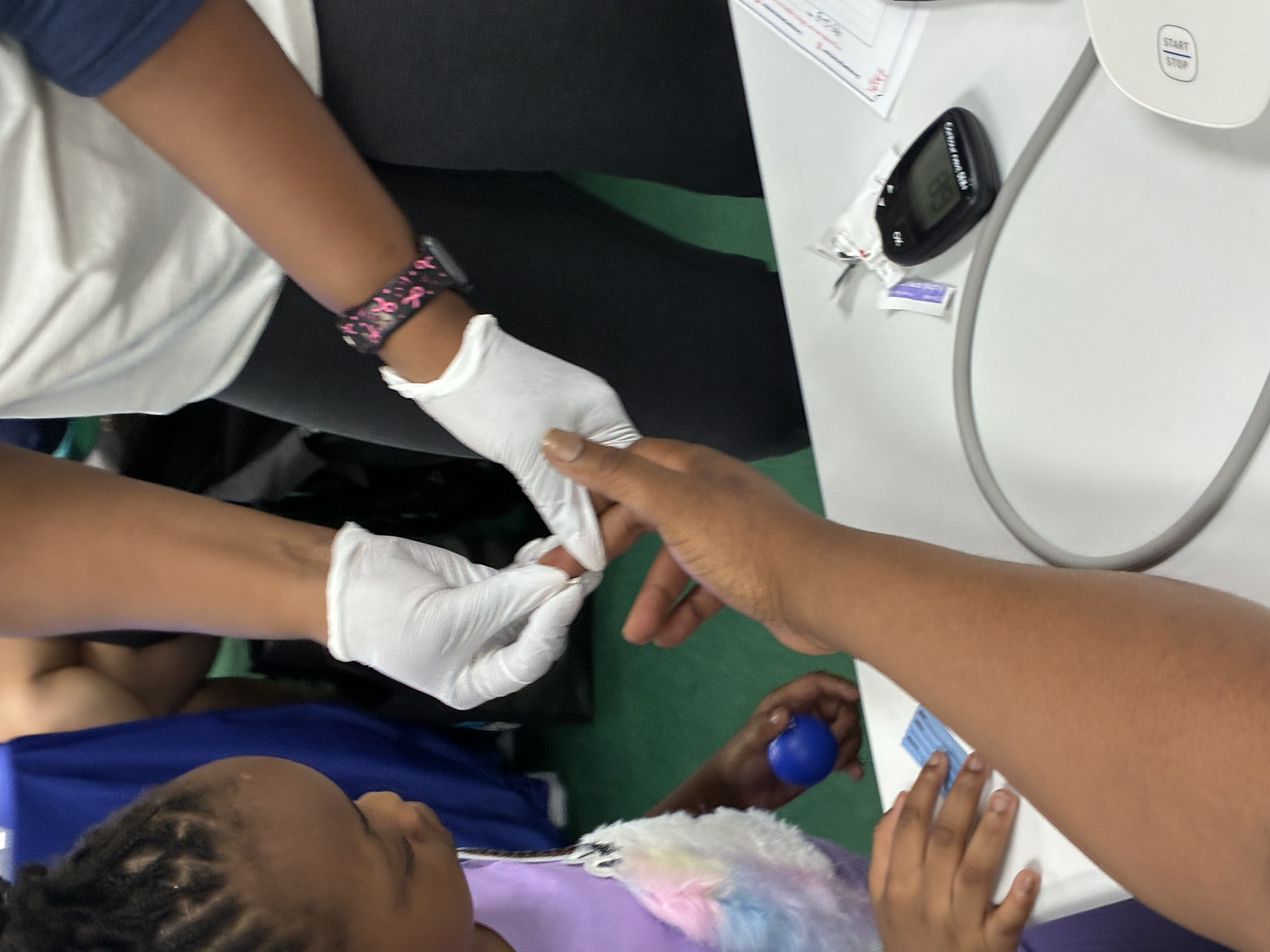 Children in blue and white shirts gathered around a white table, with some holding small blue balls and a person using a glucometer. There are papers, a monitor, and other medical items on the table, suggesting a health or medical activity in progres