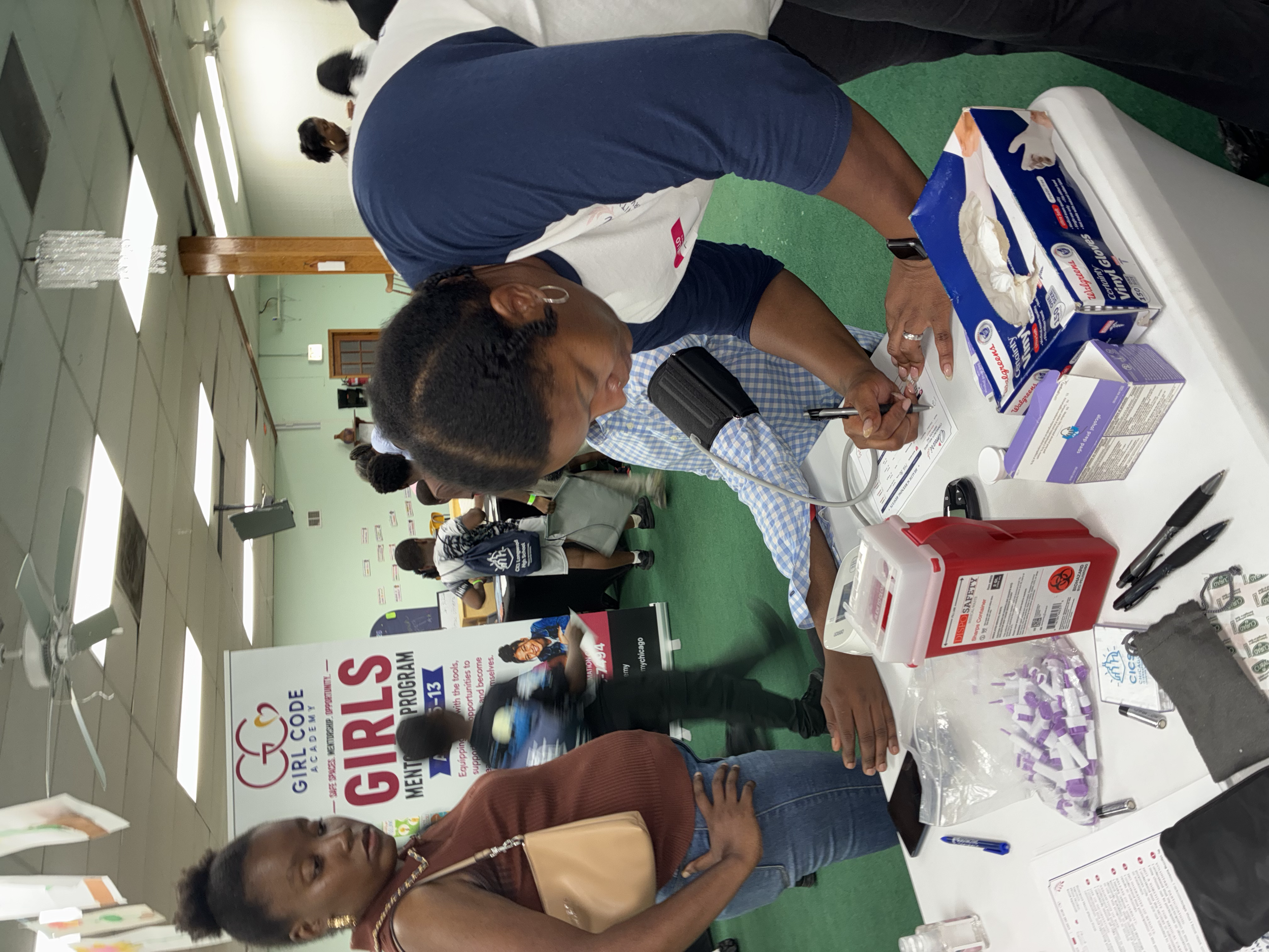 Several people at a health fair or community event, with a woman in the foreground examining brochures or pamphlets on a table that has healthcare-related items and information. There are banners promoting healthcare organizations in the background.