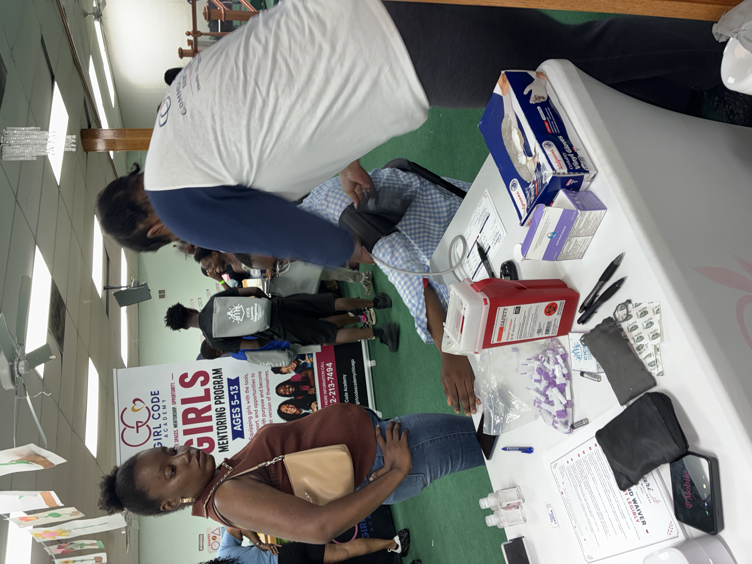 A woman at an informational booth engaging with a man, displayed on a table with brochures, pens, and small bottles of hand sanitizer. Several people are in line or browsing nearby, with banners and posters promoting a Girl Scouts event in the backgr