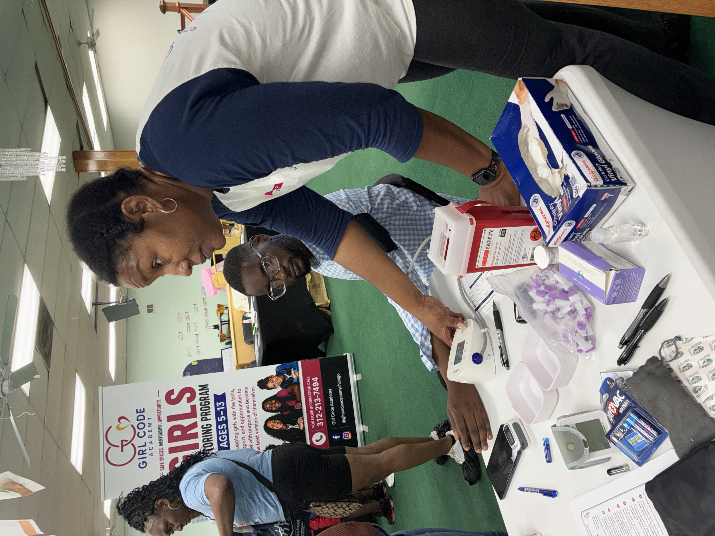 People gathered around a table at an informational event, looking at brochures and chatting, with a sign in the background promoting educational programs.