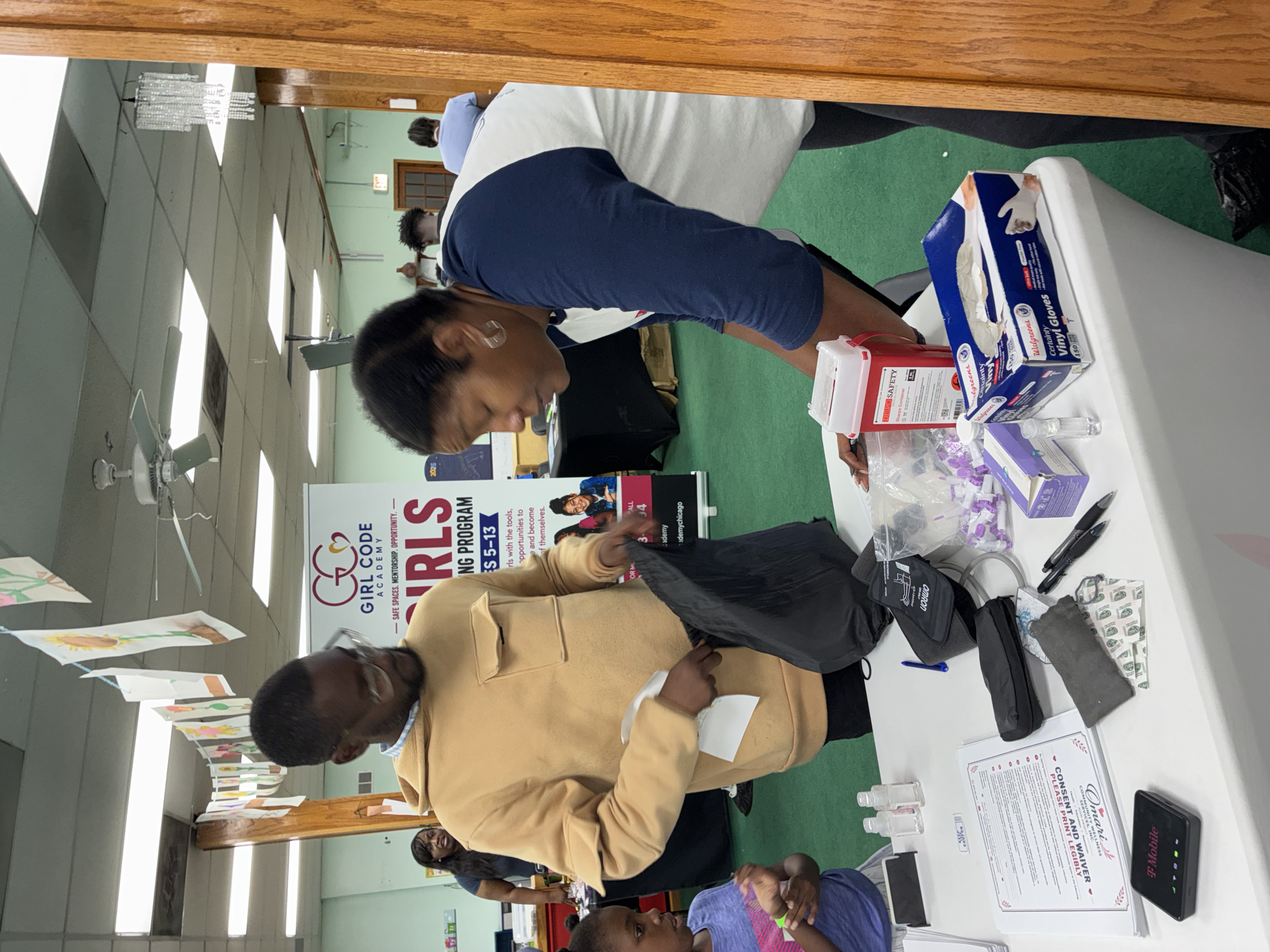 People gathered around tables at an event, with various informational materials, sign-up sheets, and promotional items on display, in an indoor setting with ceiling fans and fluorescent lighting.