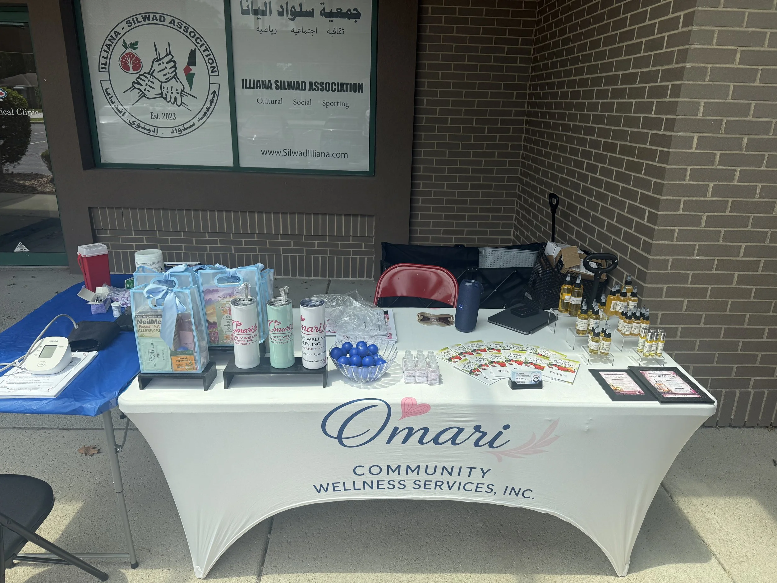 A table set up outdoors with various wellness and healthcare items for a community event, including informational pamphlets, bottled oils, and small tins, in front of a sign for the Illiana Silwad Association.