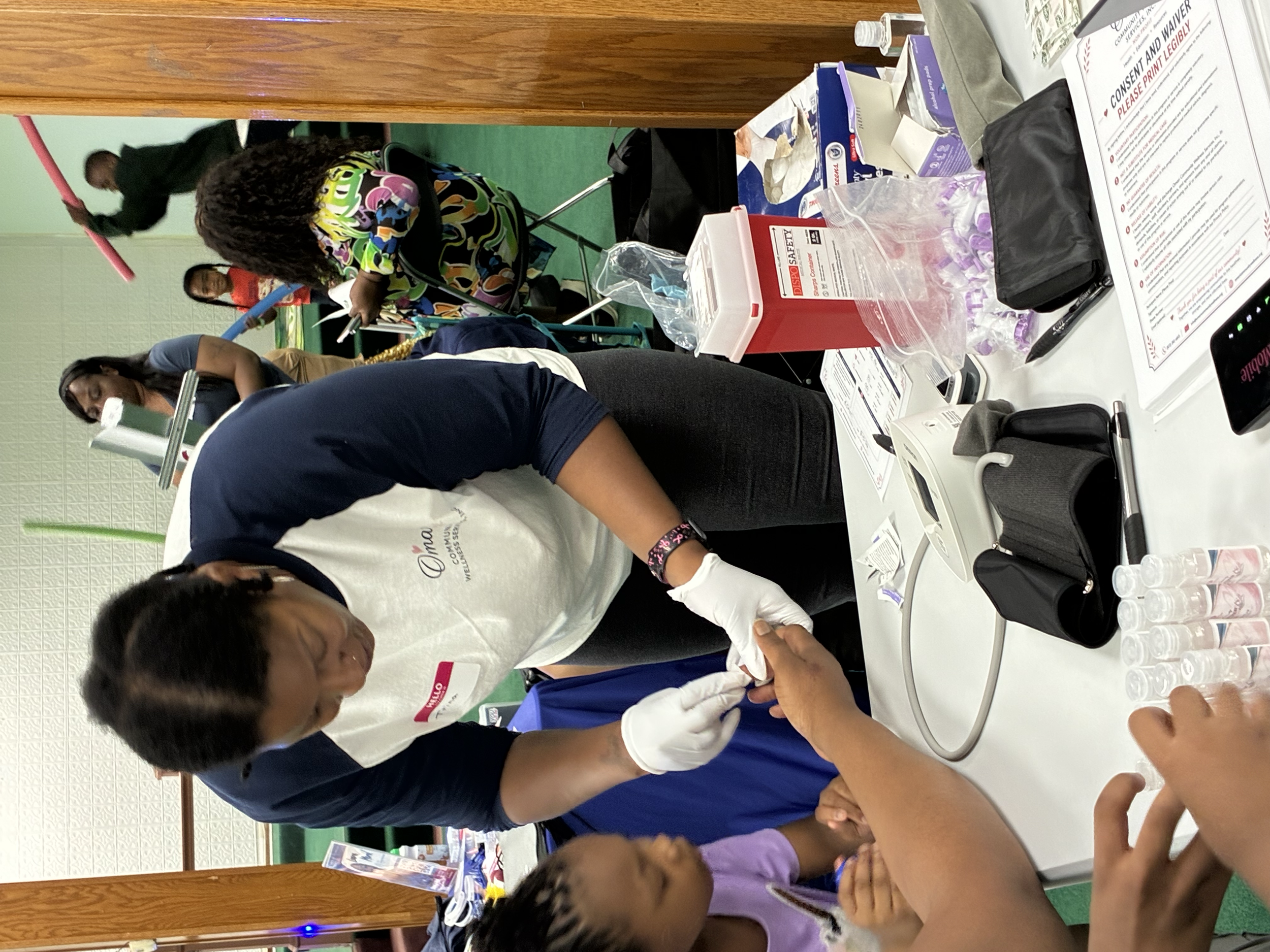 Group of children and adults engaging in an activity at a table in an indoor setting, with various supplies and informational materials visible on the table.