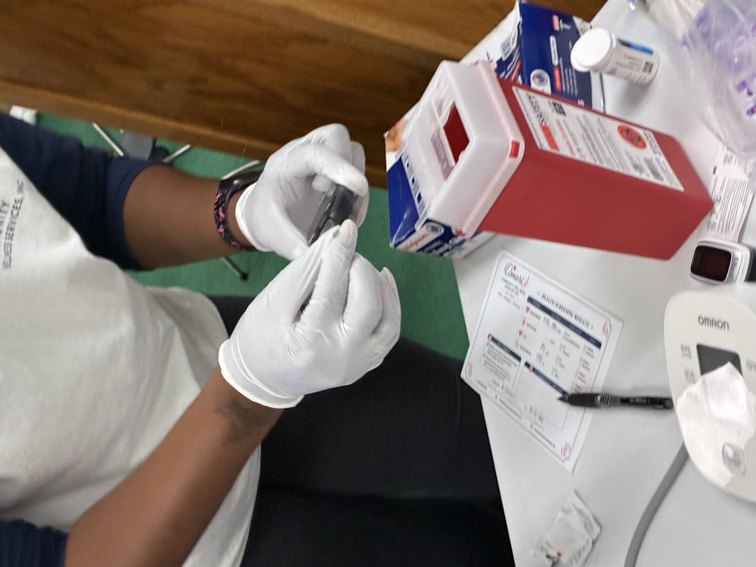 A person wearing white gloves is preparing to administer a vaccine or injection into a person's arm, which is visible in the lower part of the image. The table contains boxes of medical supplies, medication bottles, a thermometer, and informational p