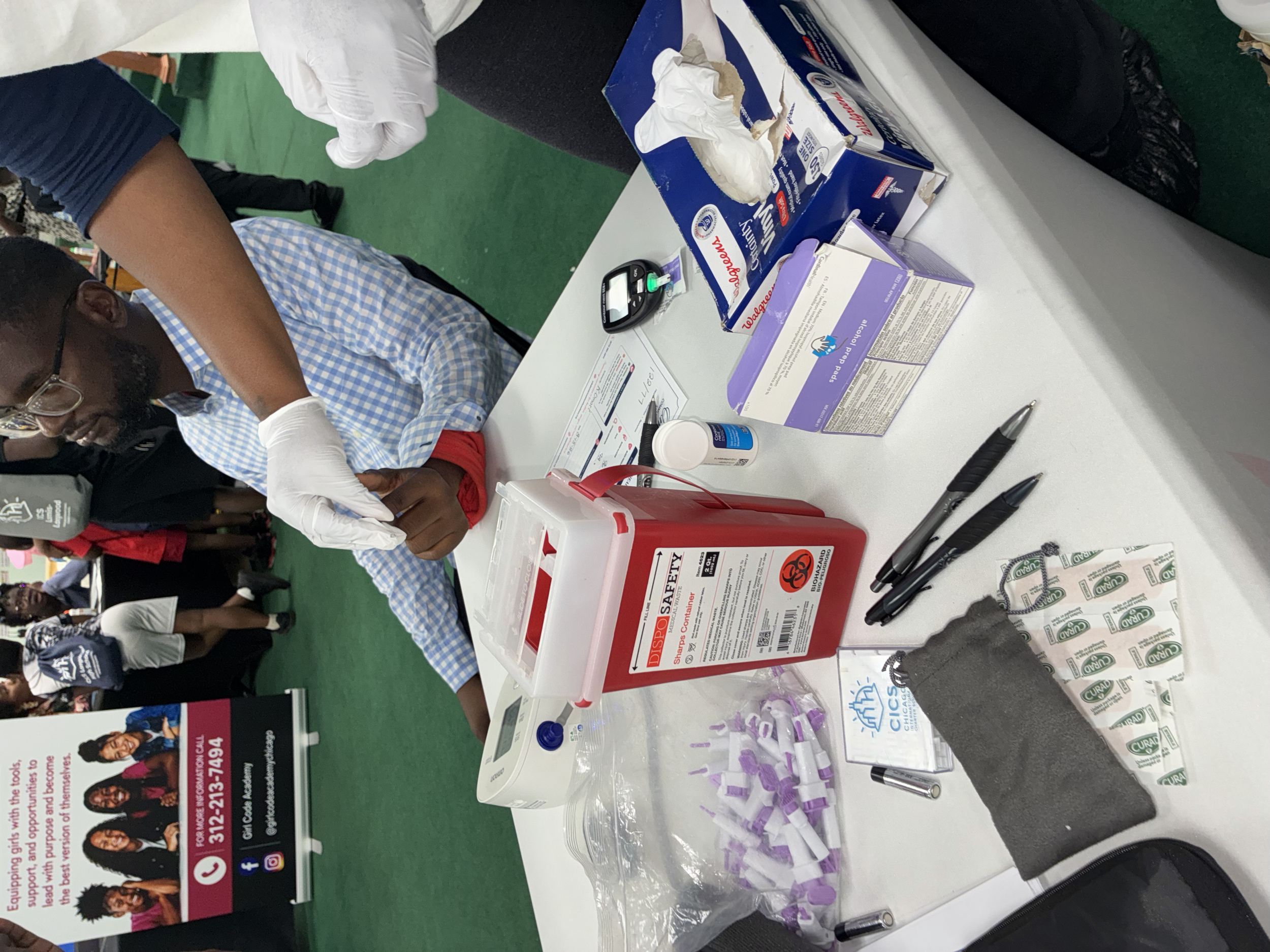 A healthcare table with medical supplies including a box of gloves, purple-capped vials, a sharps disposal container, a blood glucose meter, pens, and paper. People and informational banners are visible in the background.