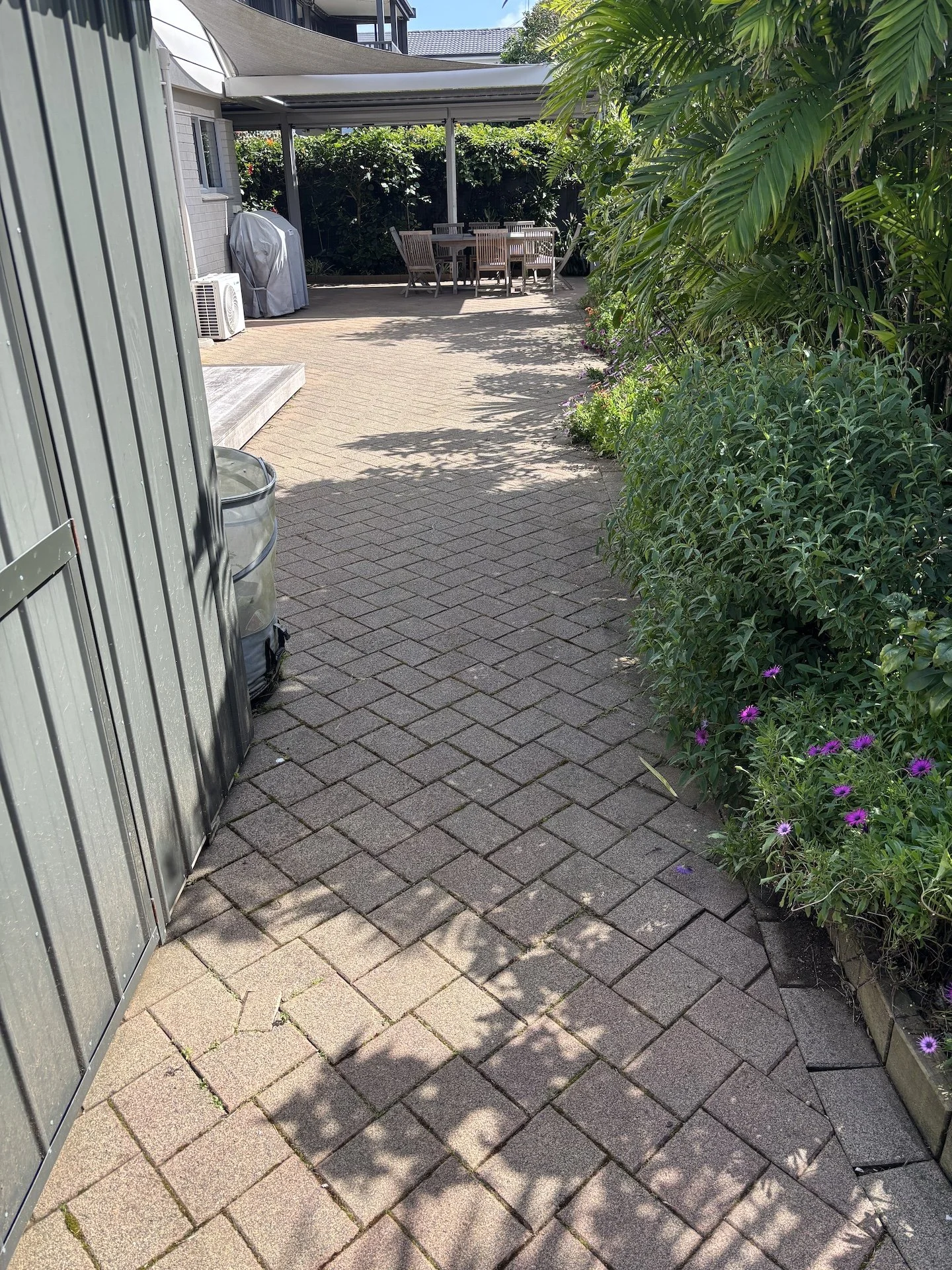 Paved backyard patio area with outdoor dining table and chairs, surrounded by lush green plants and flowers, with a shade sail overhead.