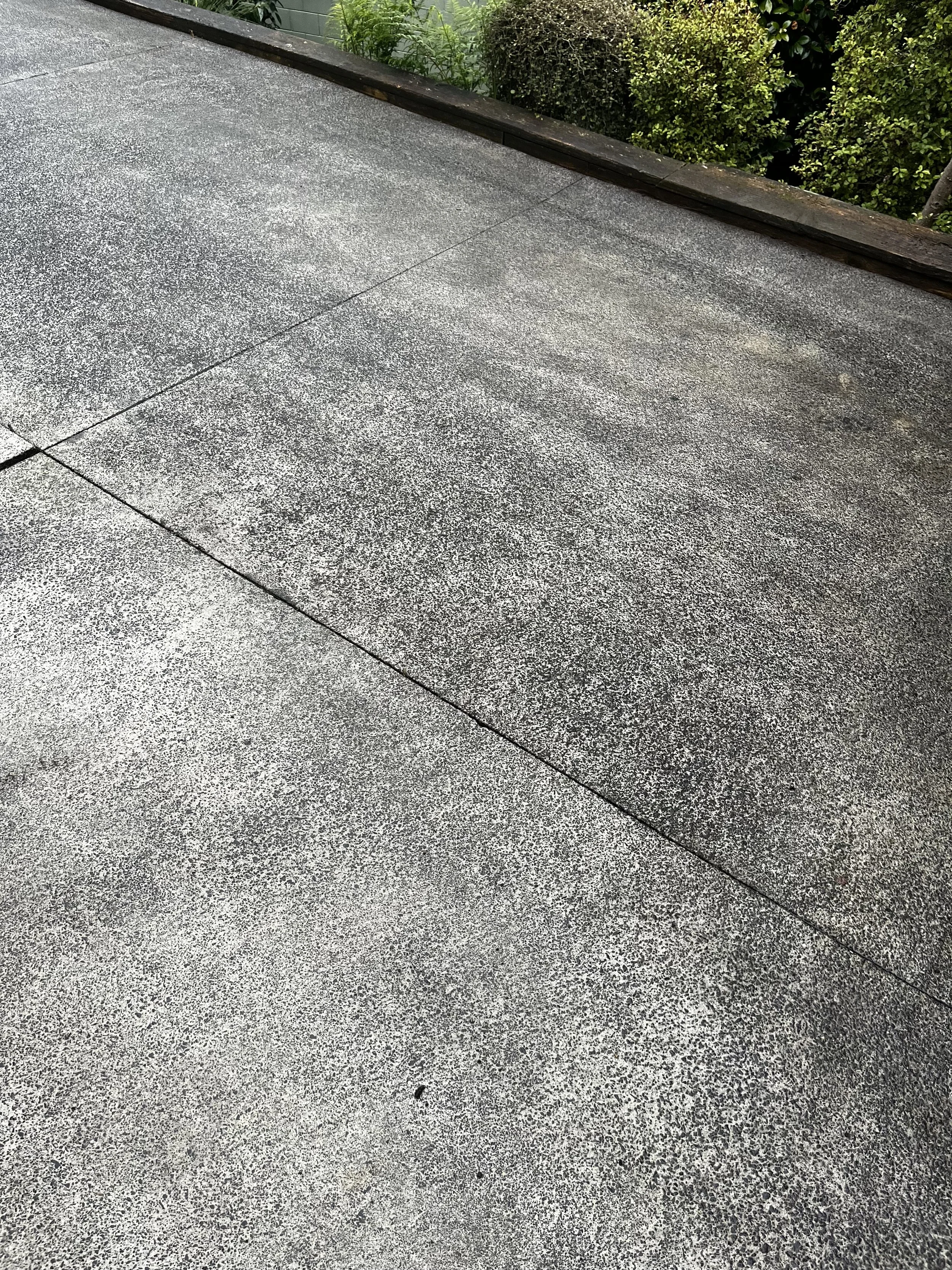 A concrete outdoor patio with a textured surface, bordered by a low wooden edge, with green bushes and plants in the background.