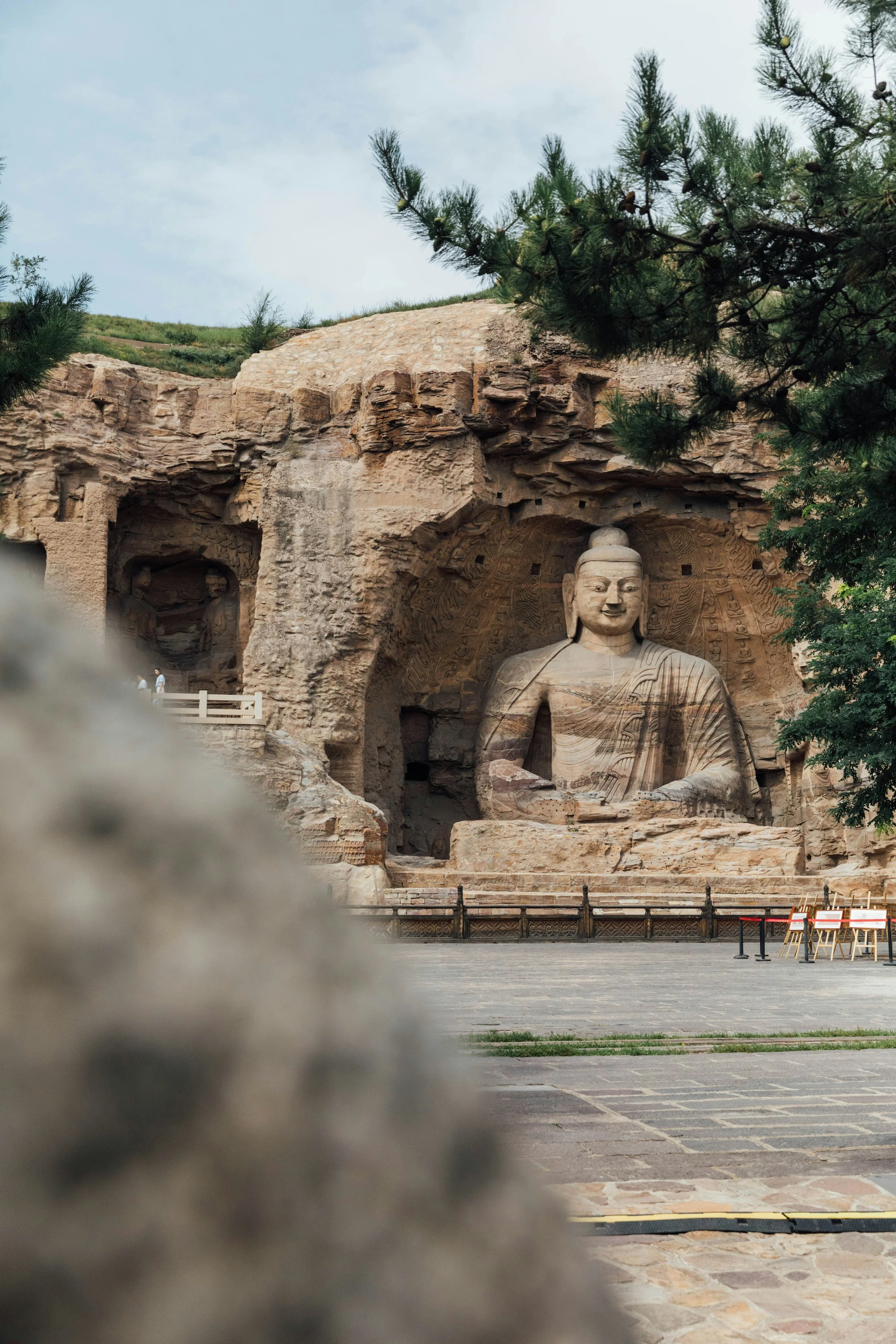 Large stone Buddha statue carved into a rock face, with trees and a cloudy sky in the background.