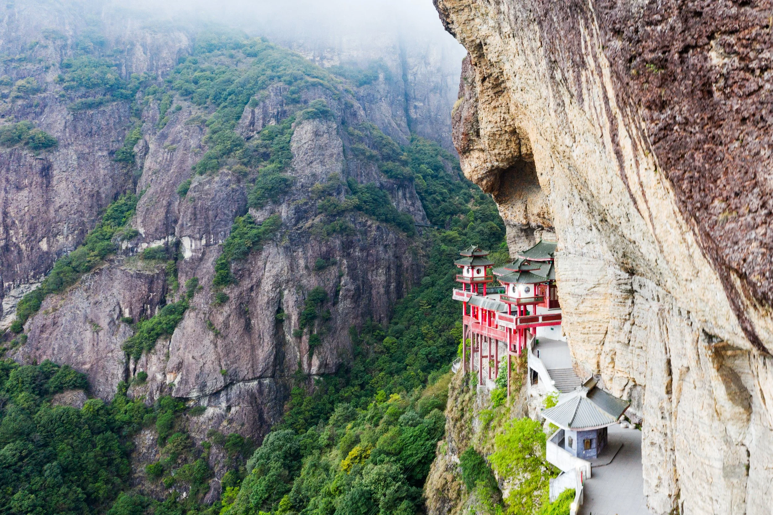 Traditional Chinese temple perched on a rocky cliffside with lush green mountains in the background.