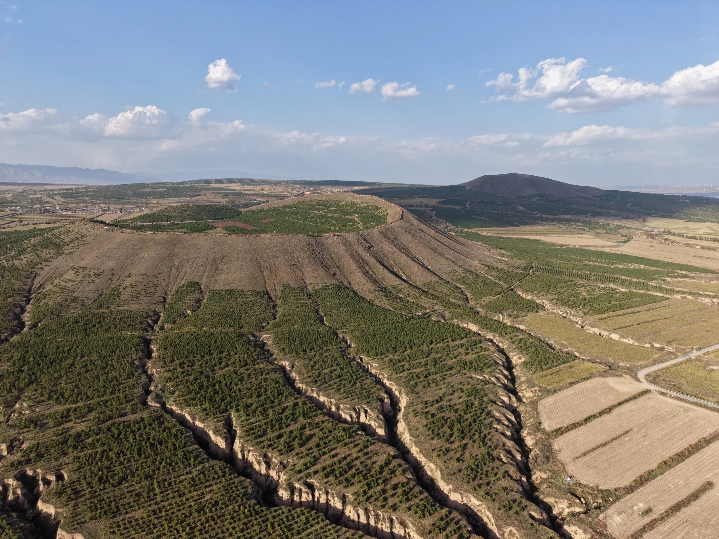 Aerial view of a terraced hillside with green vegetation, agricultural fields, and a distant mountain with cloud cover in the sky.
