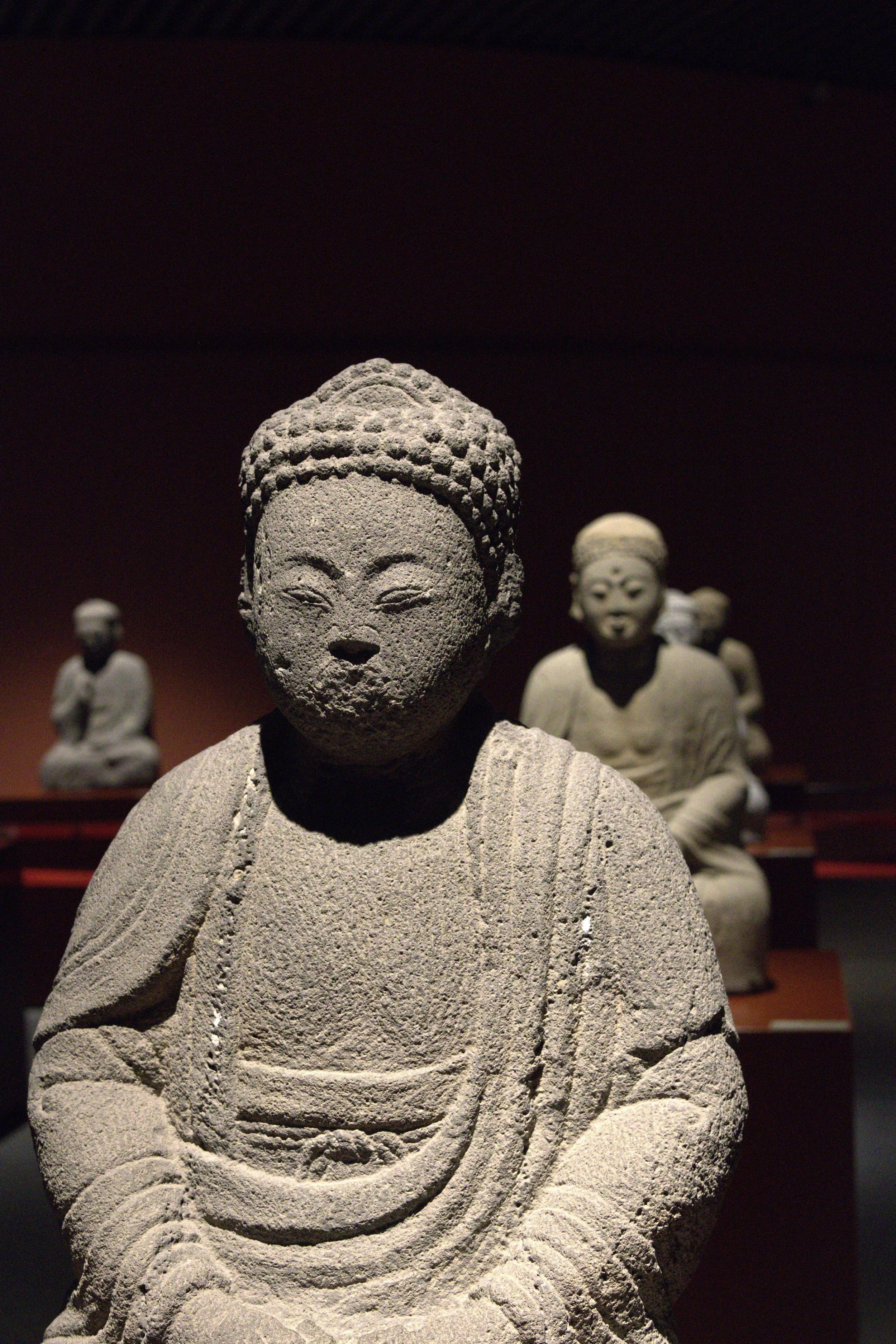 A collection of ancient stone statues depicting seated figures, with the central figure in the foreground showing a serene face, textured hair, and draped clothing, in a dimly lit museum setting.