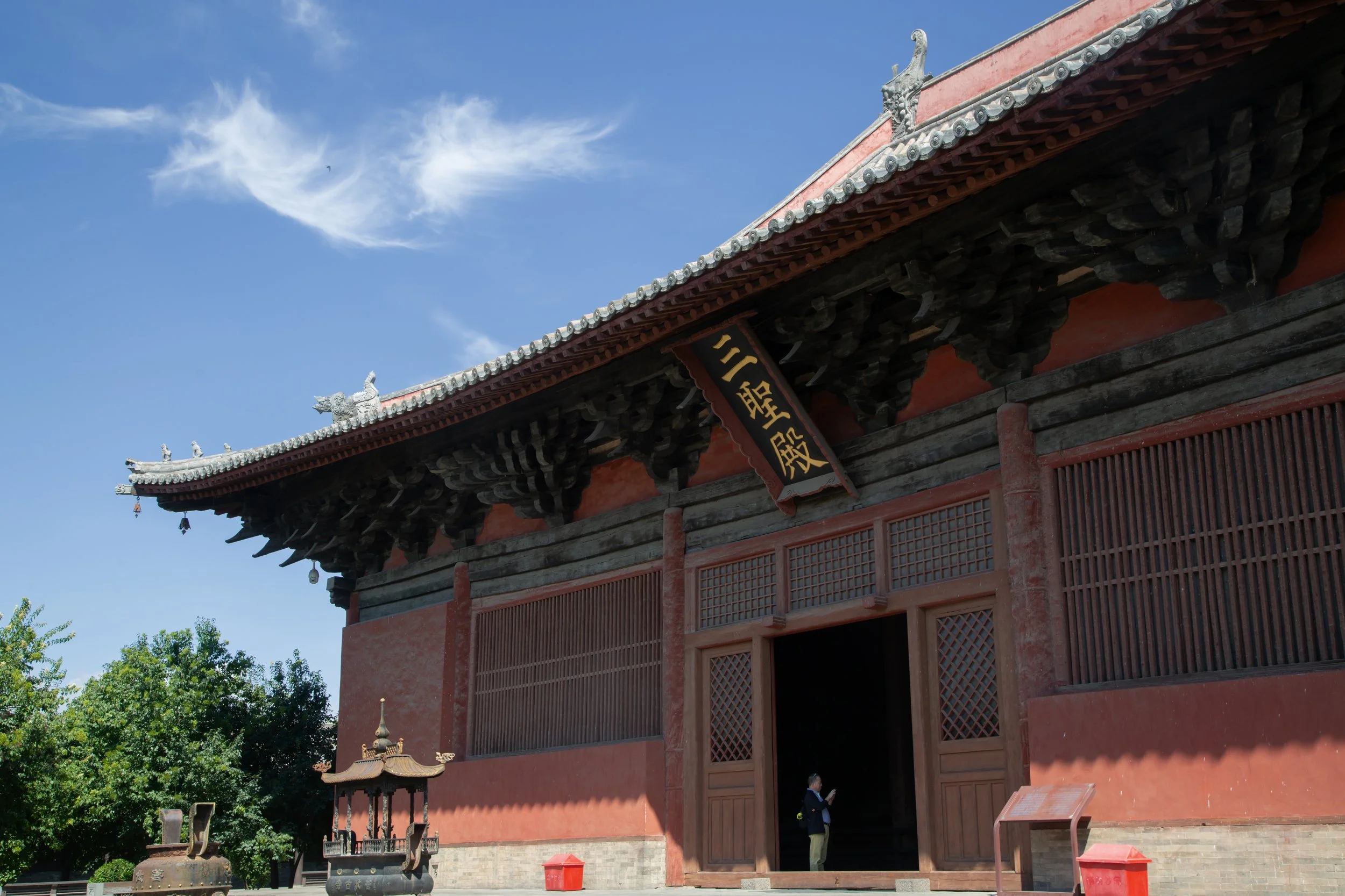 Traditional Asian temple with red walls and a decorative roof, a person standing near the entrance, a small pagoda-style structure nearby, and green trees against a blue sky with some clouds.