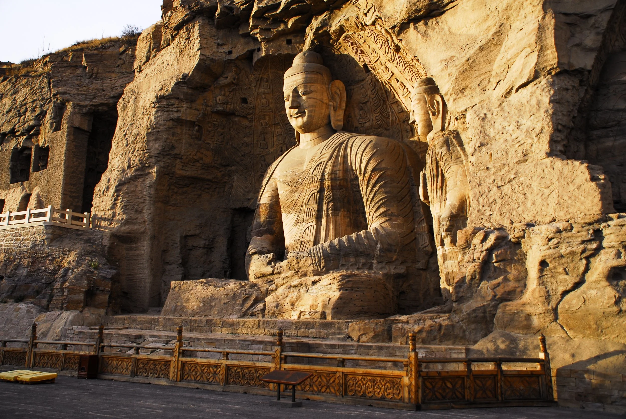 The image shows a large Buddhist sculpture of a seated Buddha carved into a rocky cliff face, with intricate details and surrounding carvings, bathed in warm sunlight.