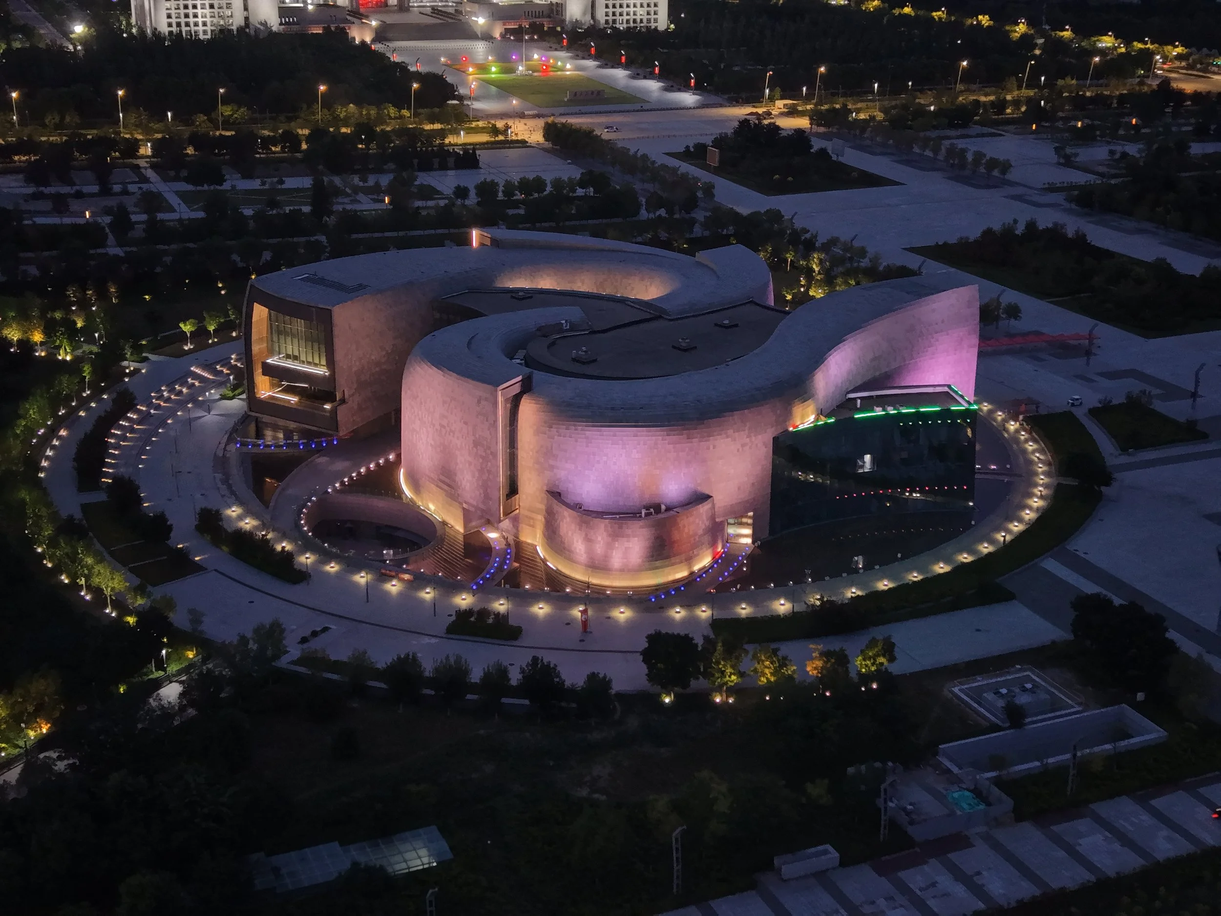 An aerial night view of a modern, illuminated building with a curved design. The building is surrounded by a circular pathway with lights, trees, and landscaped areas, with city lights and buildings in the background.